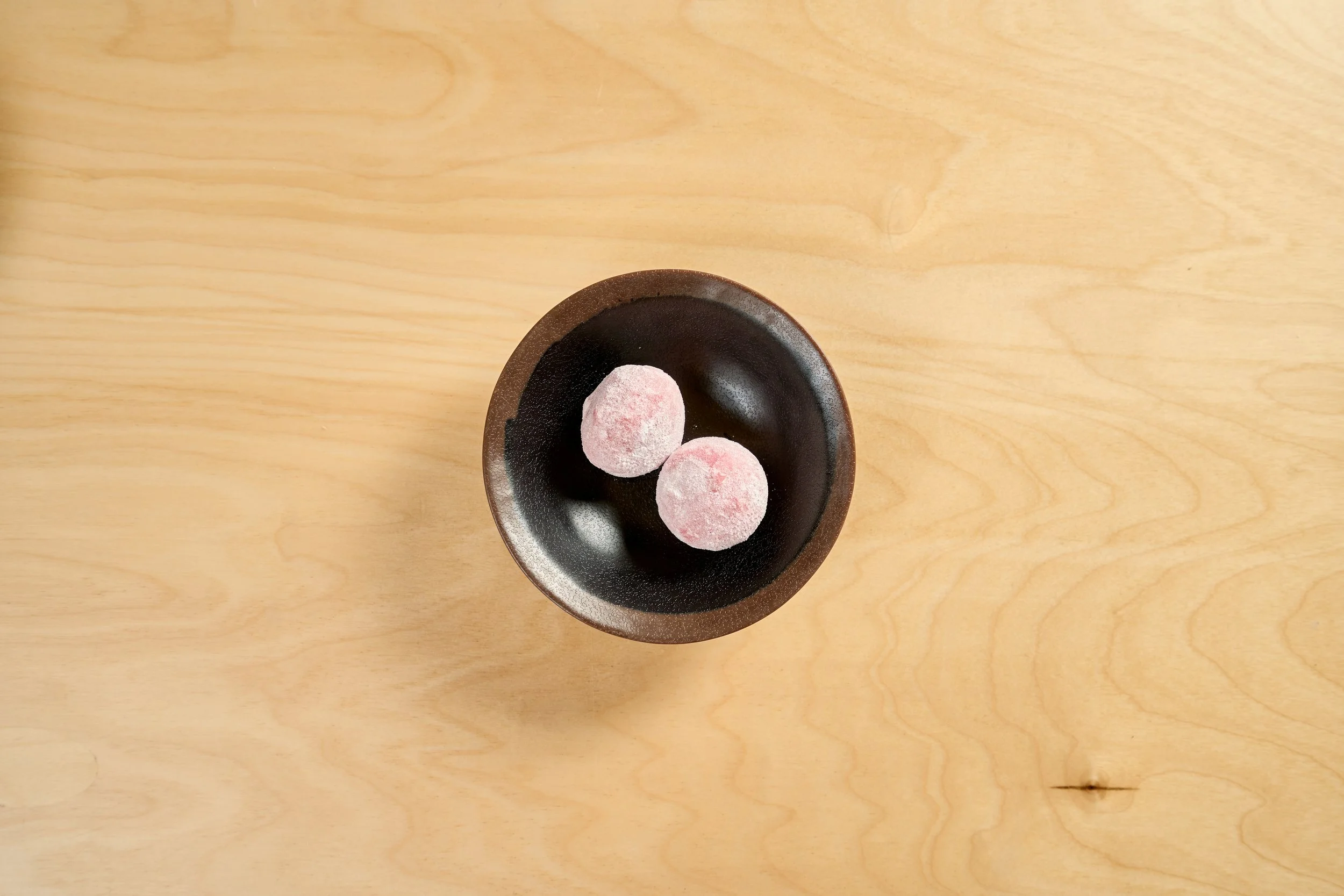 Dois doces redondos e macios, com uma camada de farinha de açúcar, em uma tigela escura sobre uma mesa de madeira clara.