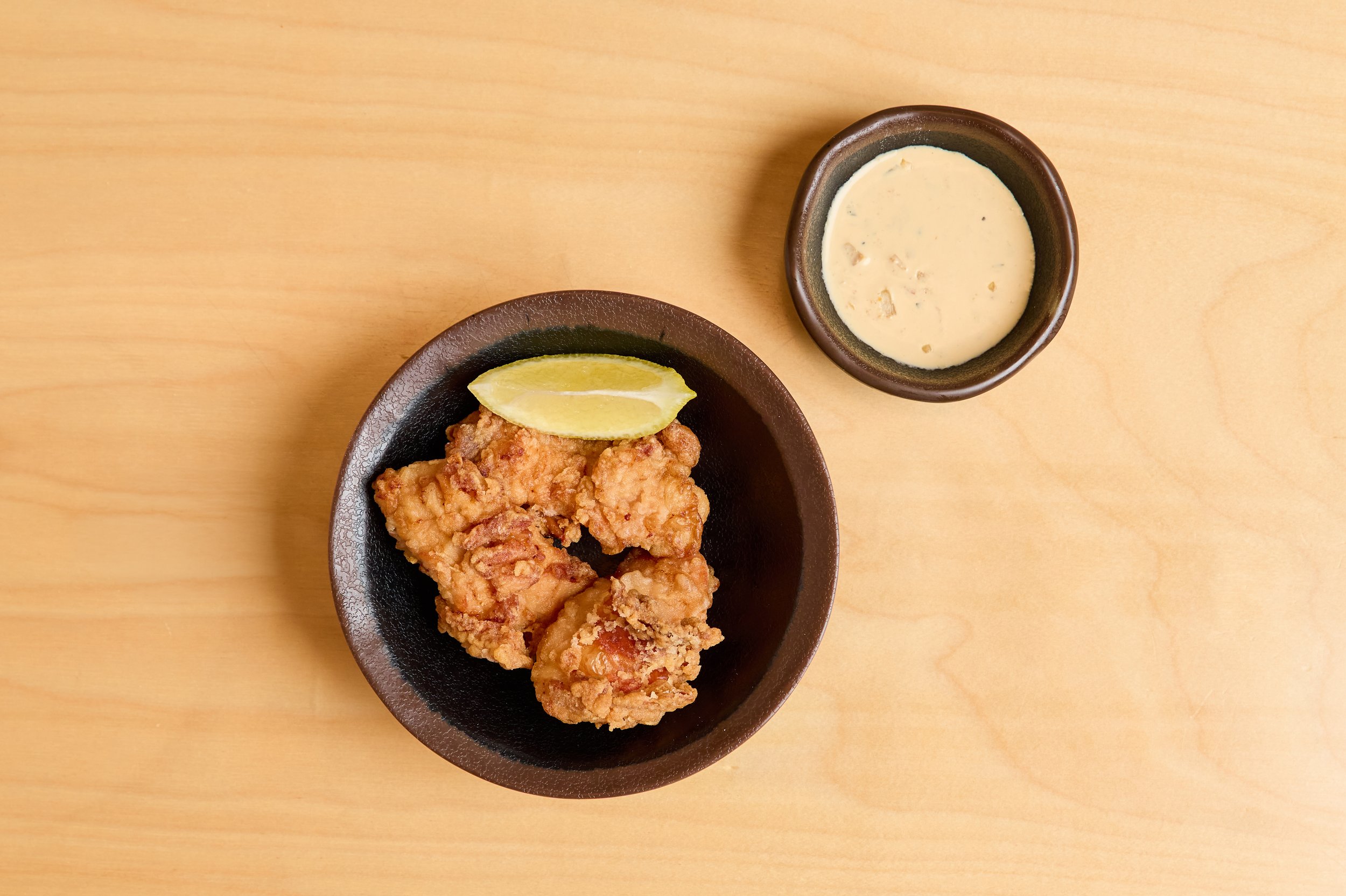 Porção de frango frito crocante com uma fatia de limão ao lado, servido em uma tigela preta, ao lado de um pote com molho branco em uma mesa de madeira clara.
