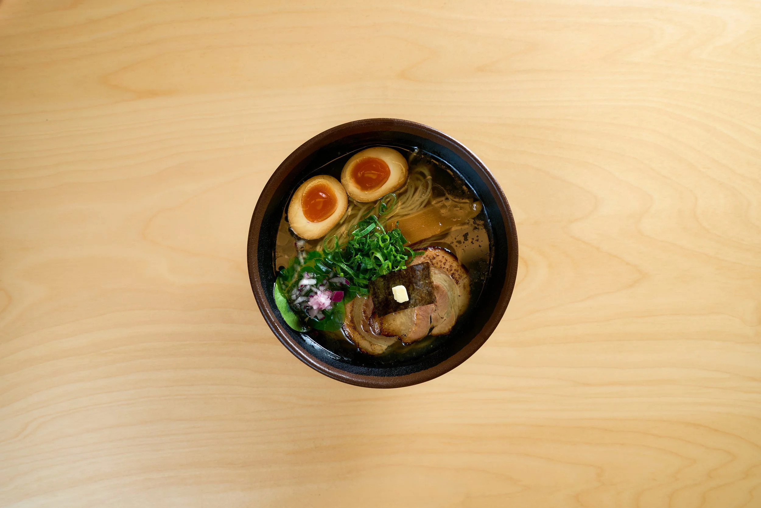 Tigela de ramen com ovo half-cocido, fatias de carne, cebolinhas, algas e outros ingredientes, sobre mesa de madeira.
