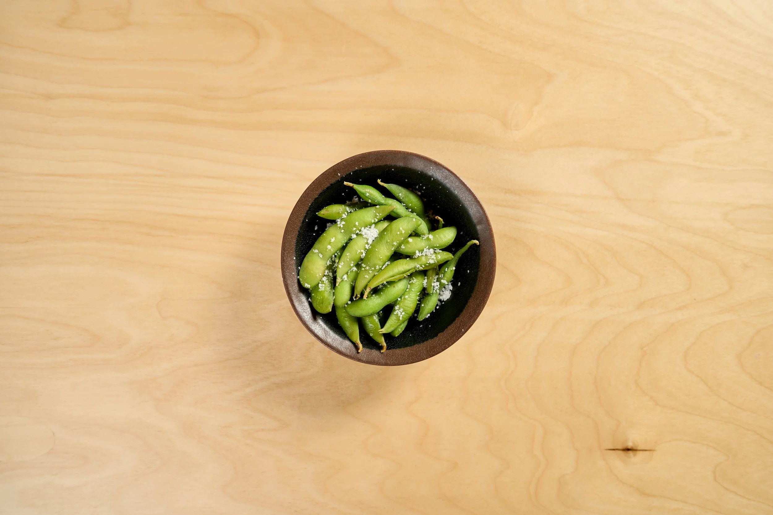 Tigelas de edamame salpicadas com sal, colocadas sobre uma mesa de madeira clara.