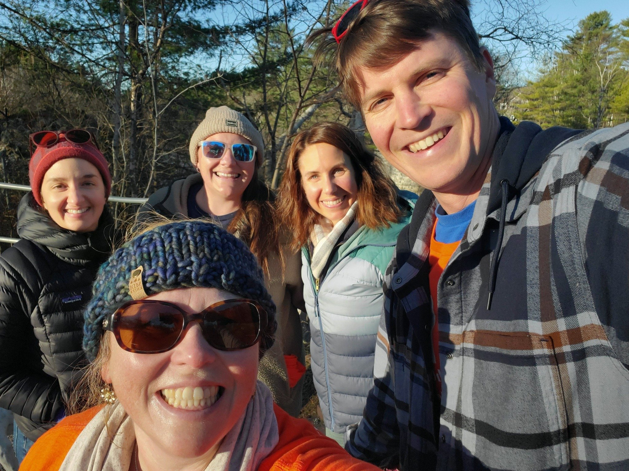 Group of five smiling friends taking a sunny outdoor selfie in winter clothing.