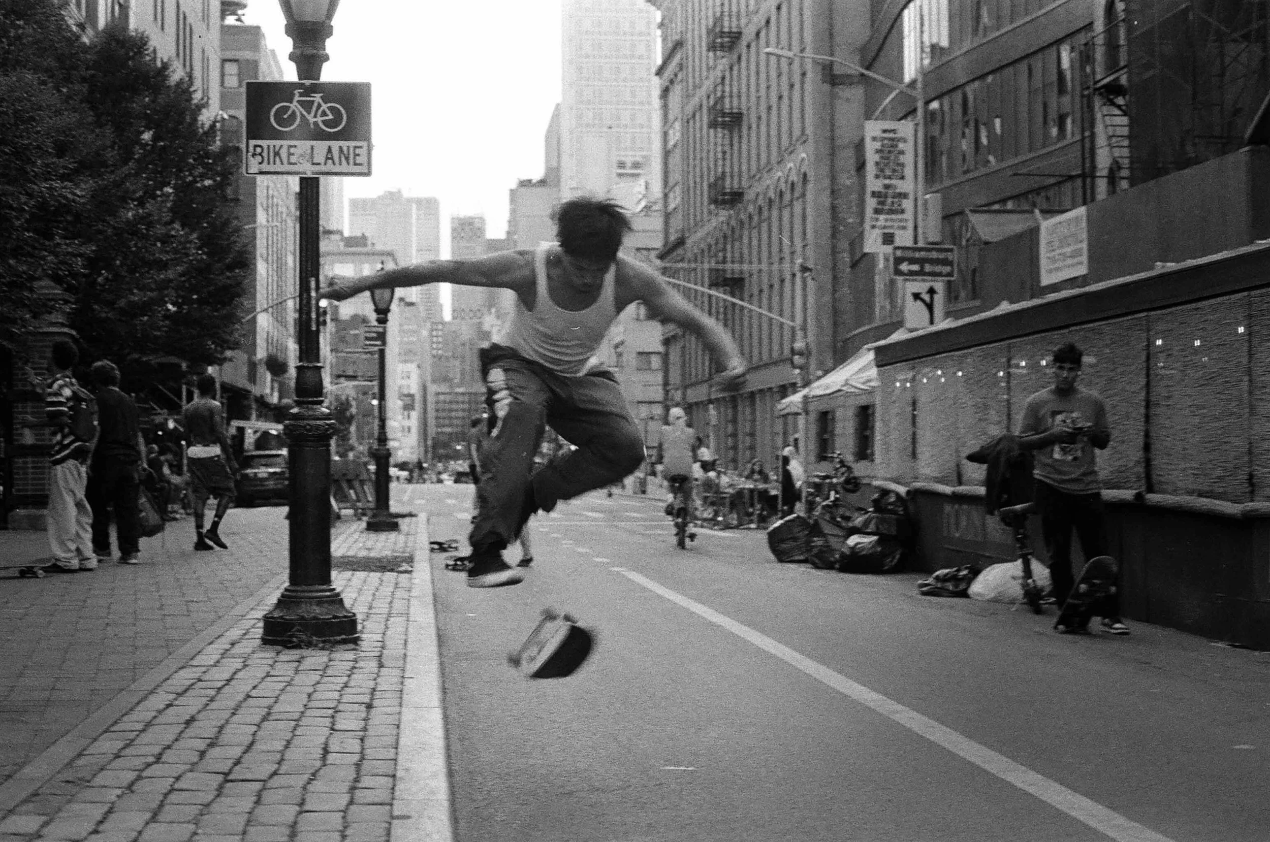 A person skateboarding on a city street with skateboard mid-air, surrounded by onlookers and tall buildings, with a sign indicating a bike lane.
