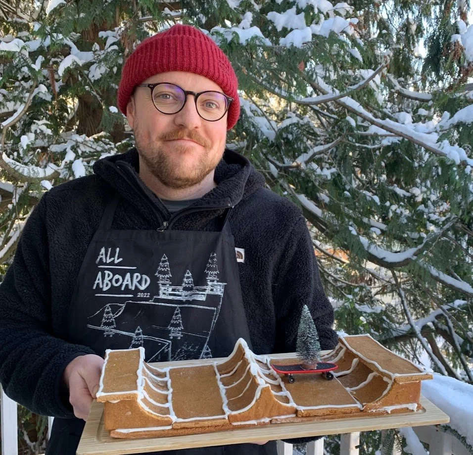 A man wearing glasses, a red beanie, and a black jacket holding a skateboard-themed gingerbread creation with white icing, a tiny tree, and snow on it, outdoors in a snowy forest.