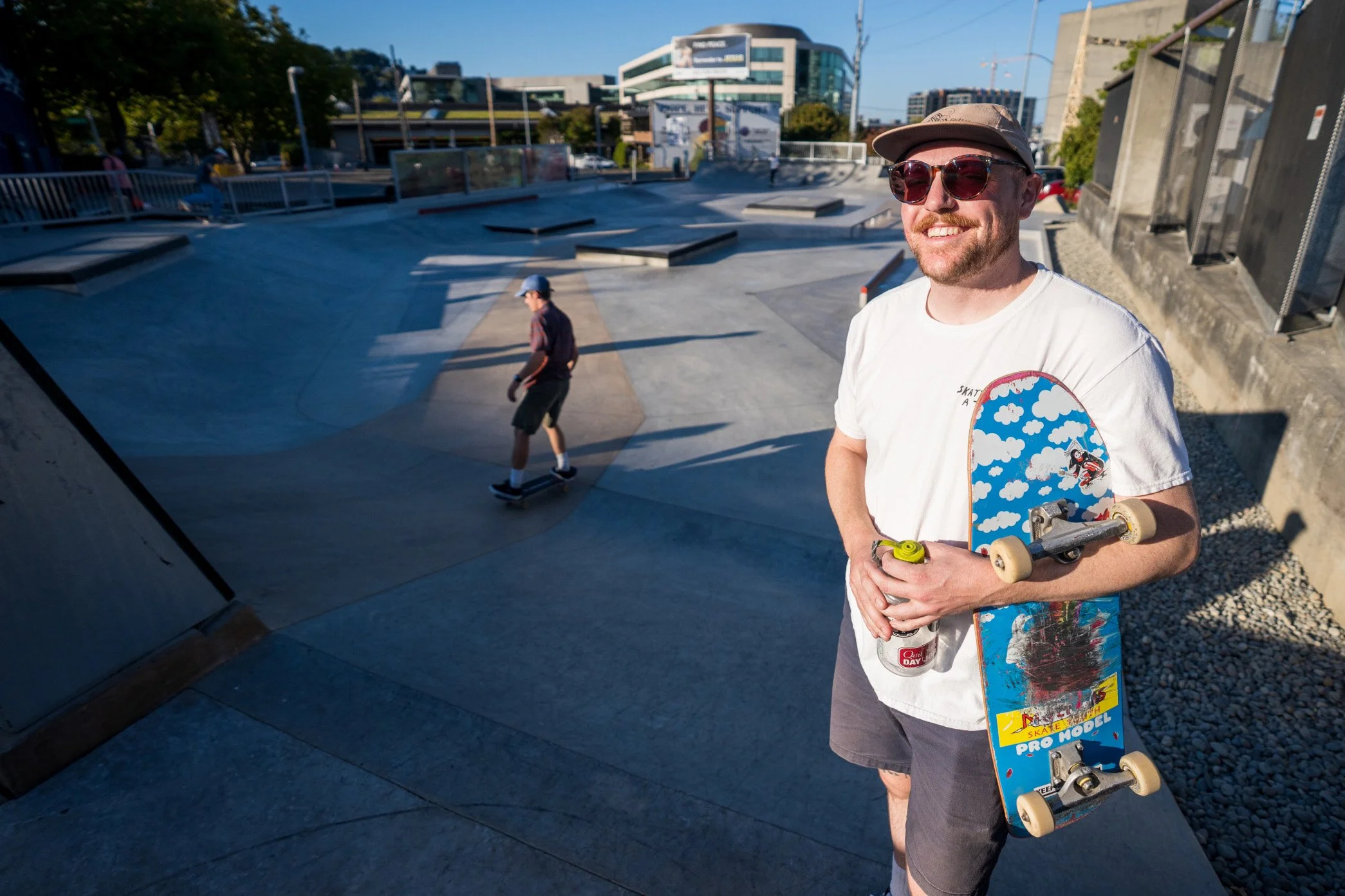 A young man with a beard wearing sunglasses and a cap, smiling while holding a skateboard with a colorful design at a skate park. In the background, another person is skating in the concrete ramps of the skate park with modern buildings and blue sky above.
