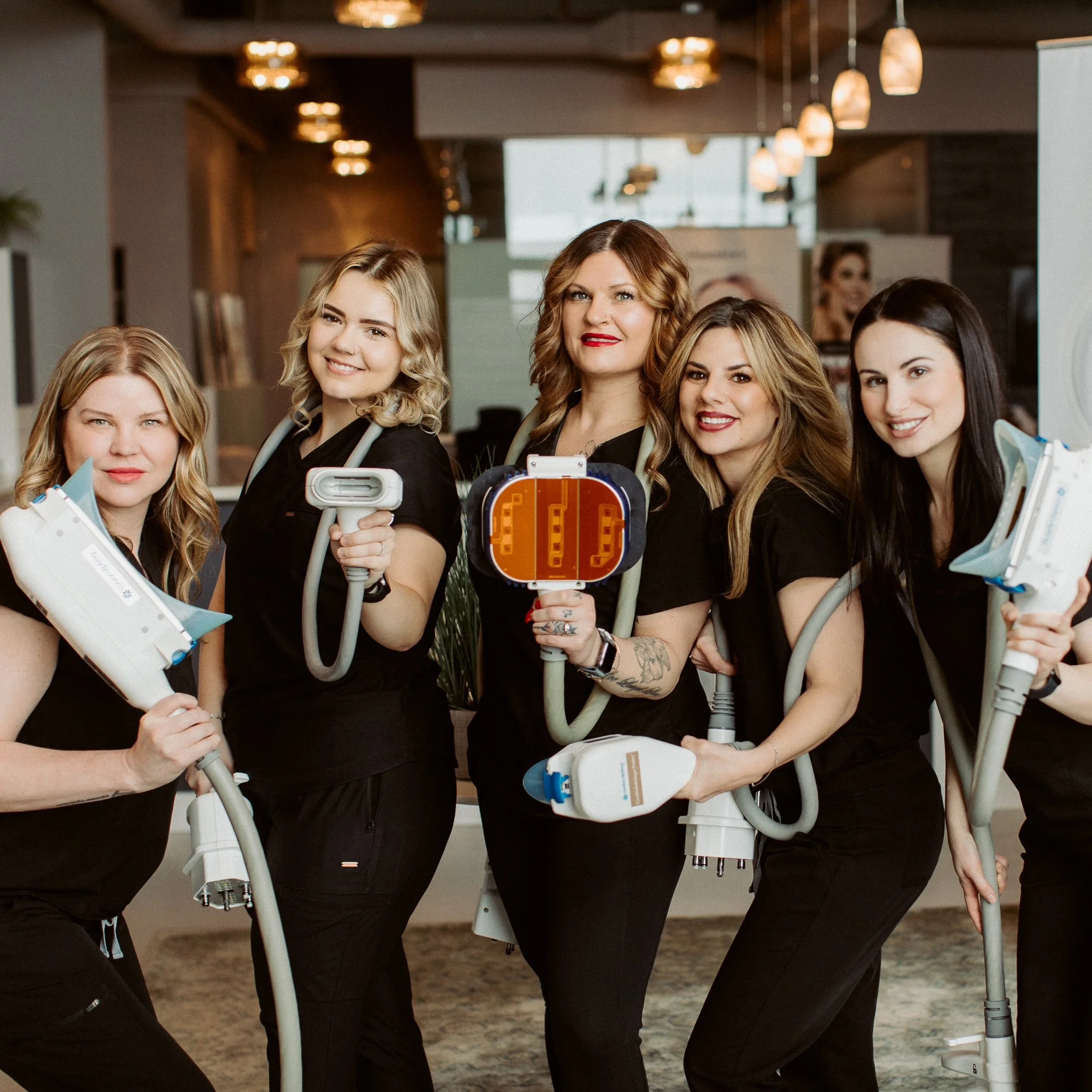 Group of women in black scrubs smiling behind medical ultrasound devices in a clinical setting.
