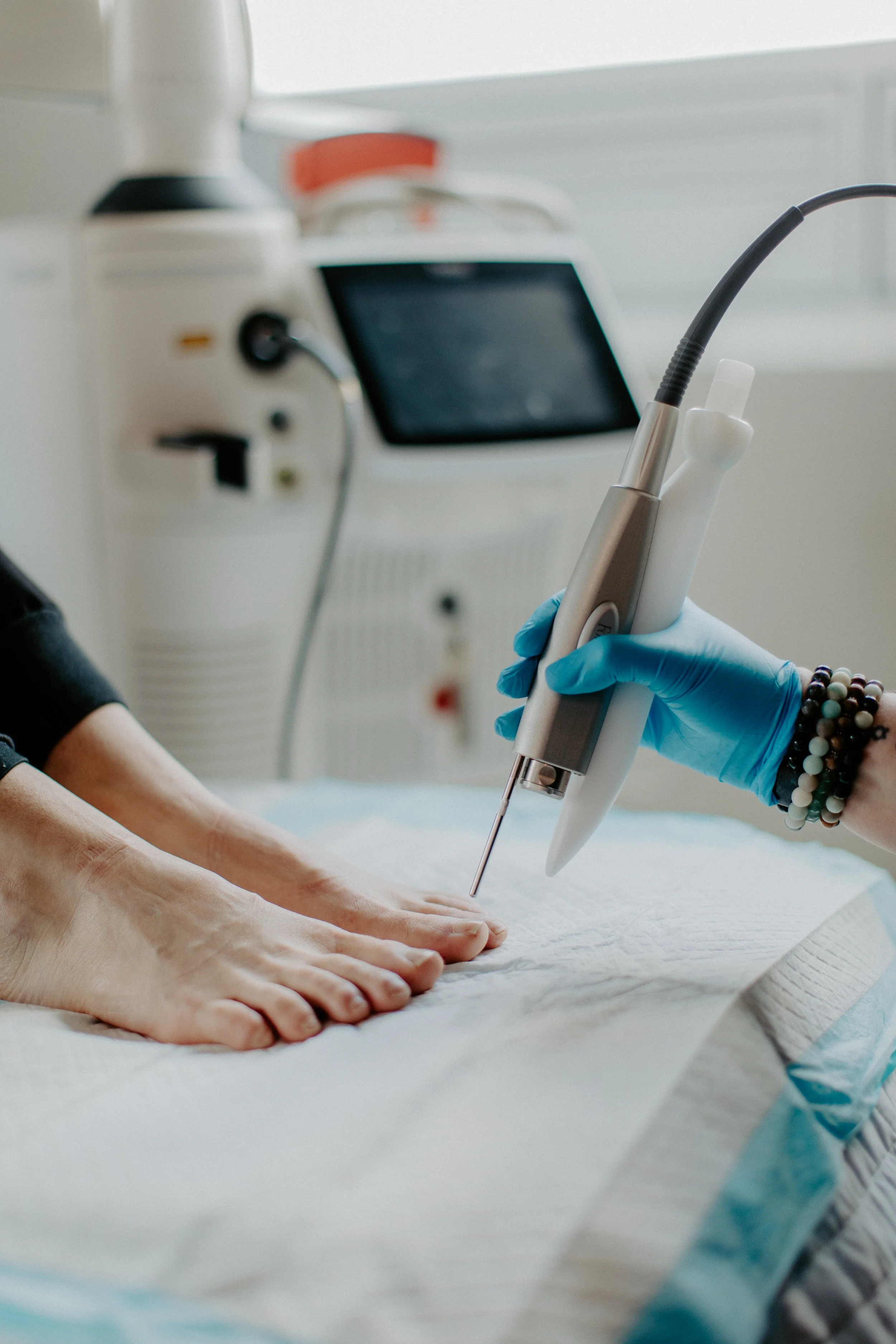 A person's hand is on a medical bed while a healthcare provider uses a handheld ultrasound device on the patient's hand, with medical equipment in the background.