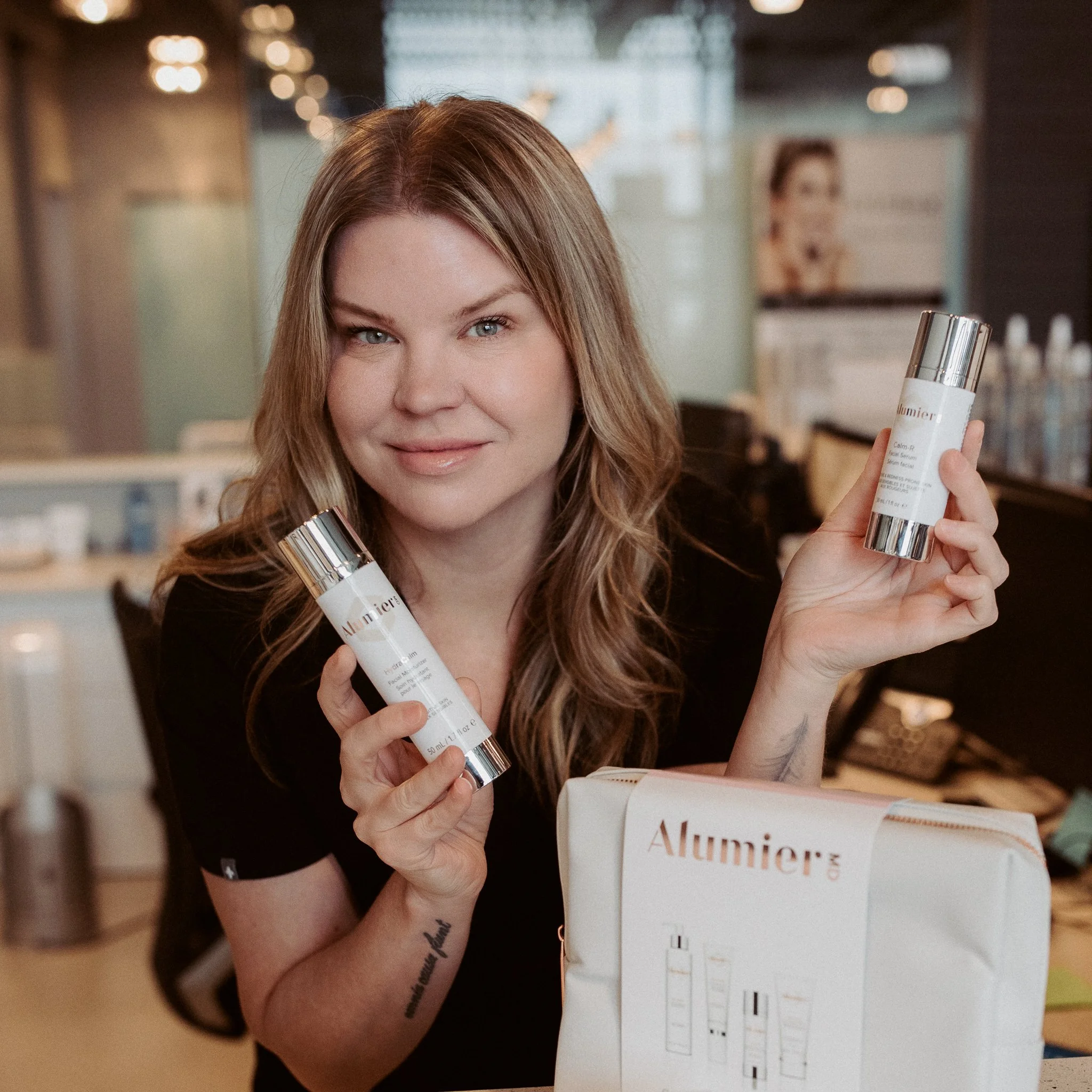 A woman with long, wavy brown hair smiling and holding two bottles of skincare products, with a box labeled 'Alumier' on the table in front of her.