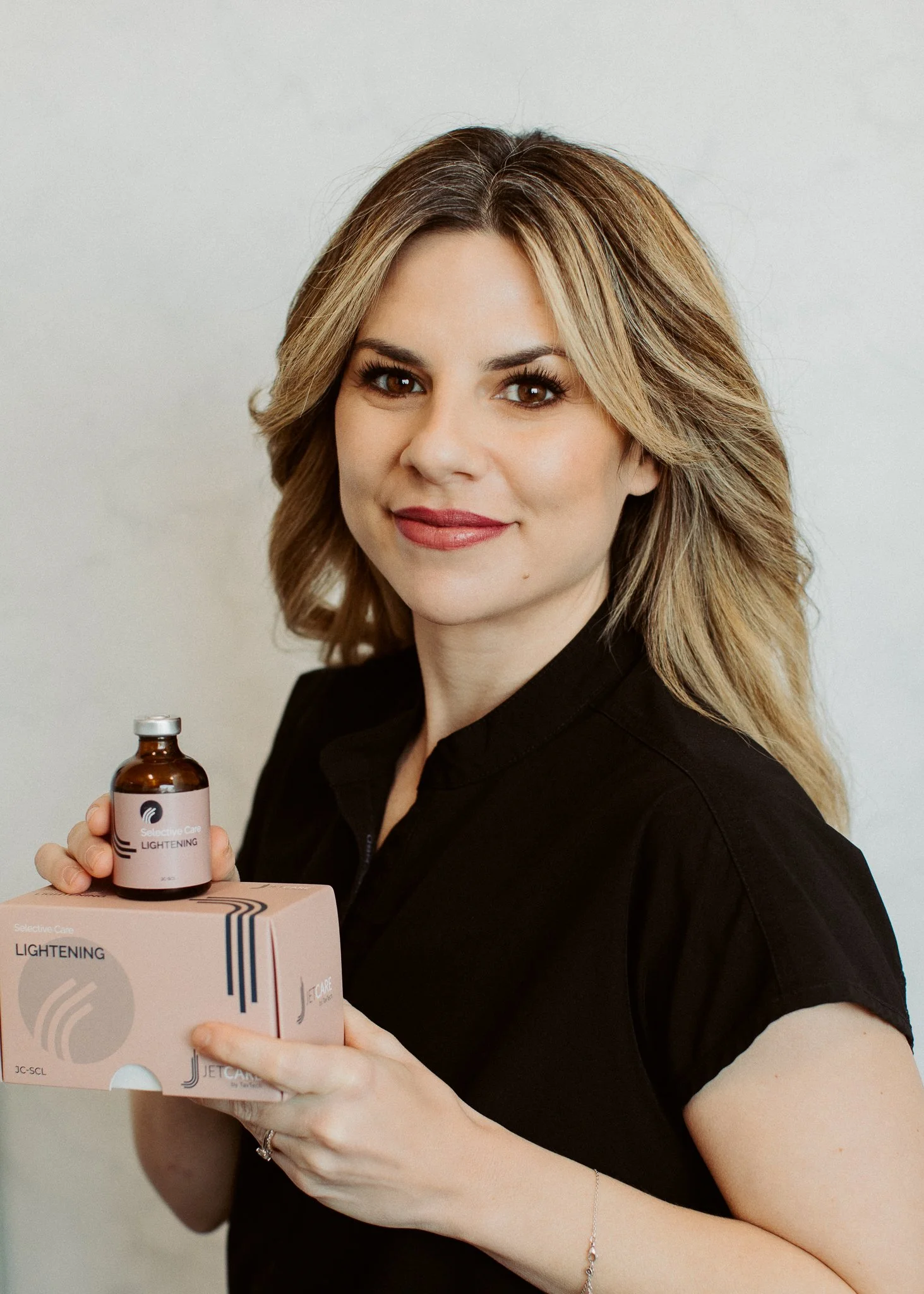 Woman with blonde hair smiling, wearing a black shirt, holding a small brown bottle labeled 'LIGHTENING' and a pink box with similar branding.