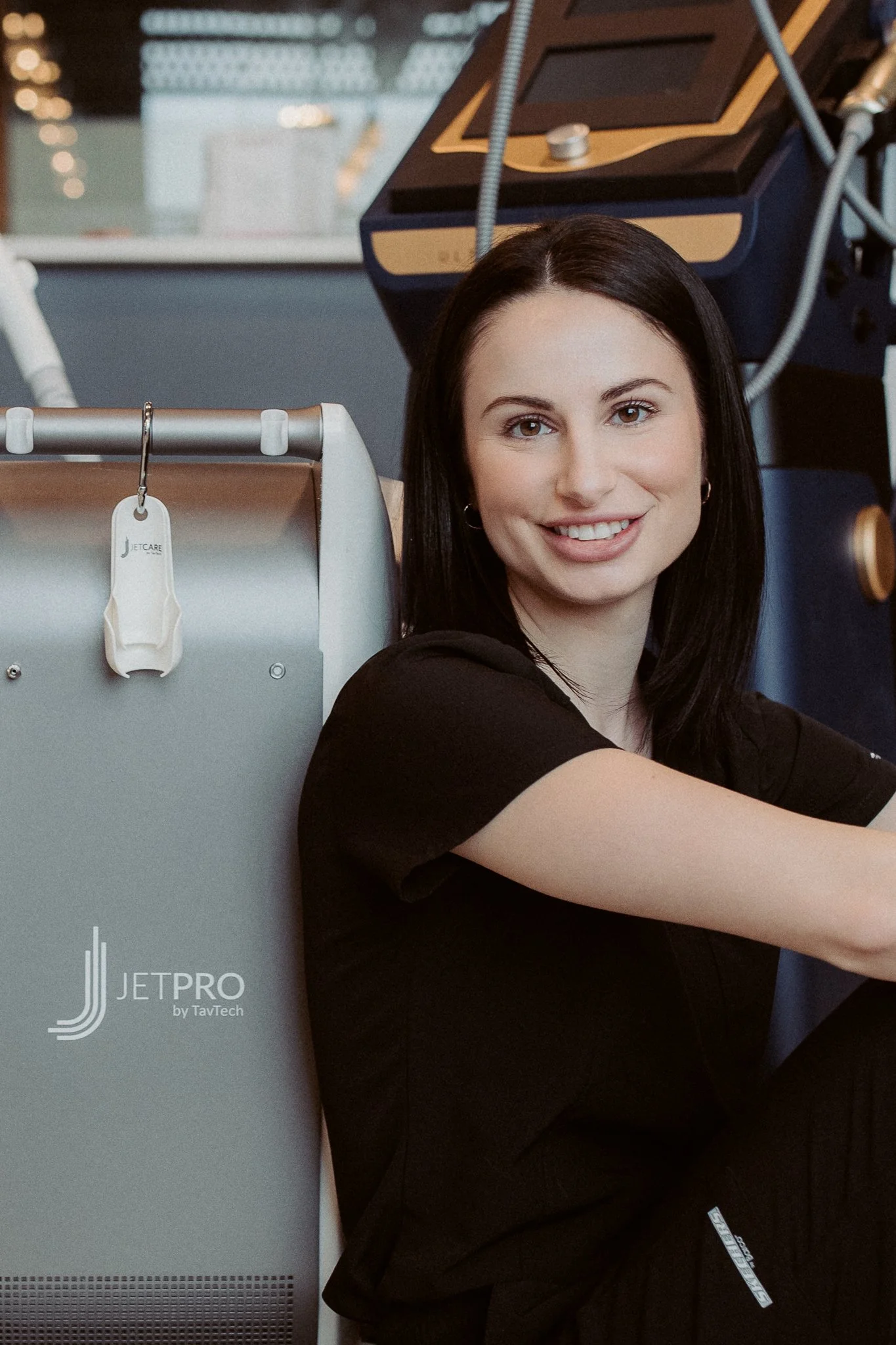 A smiling woman with dark hair sitting next to medical or aerospace equipment in a laboratory or technical setting.