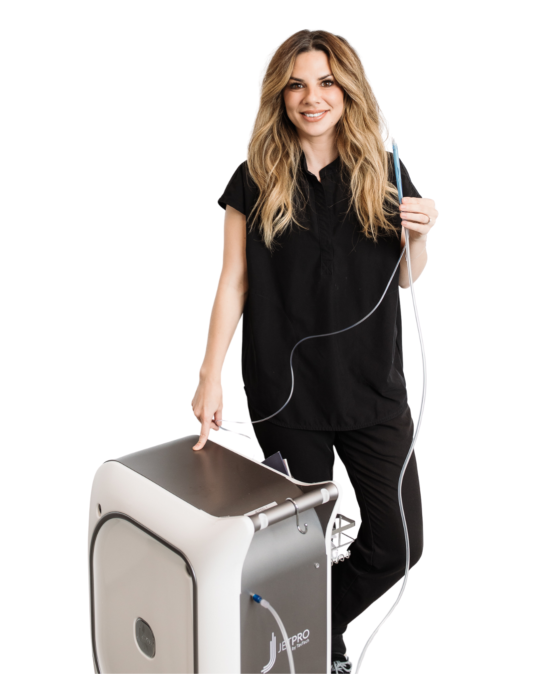 A woman with long, wavy blonde hair smiling while standing next to a medical device, pointing to it with her right hand, and holding a thin tube connector with her left hand.