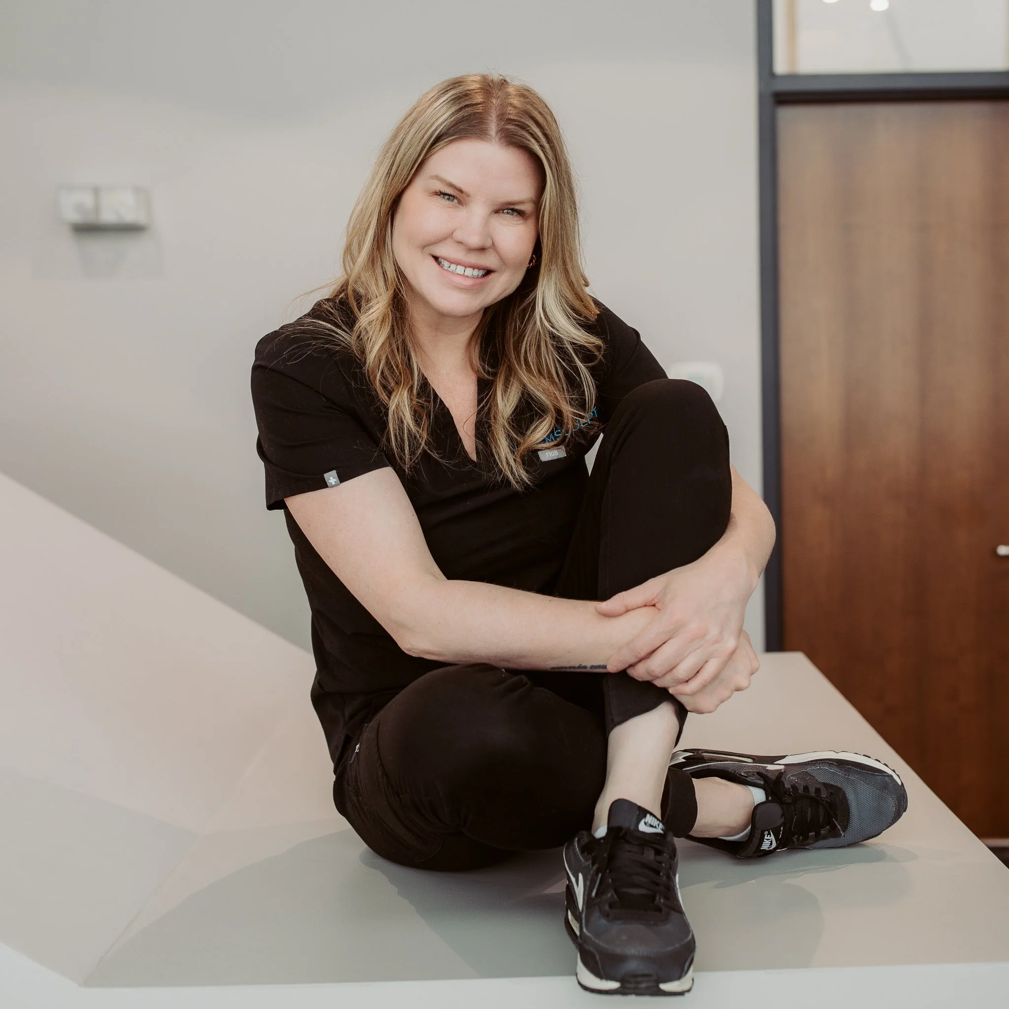 Smiling woman in black medical scrubs sitting cross-legged on a table indoors