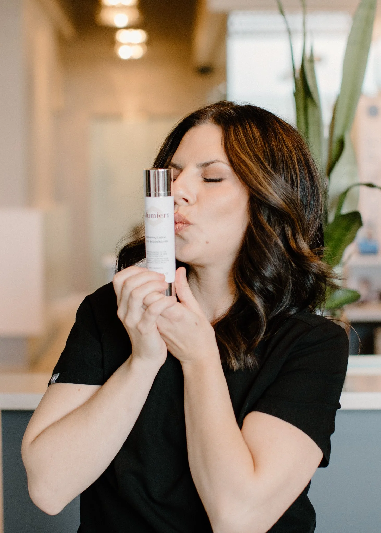 A woman with dark brown hair, wearing a black shirt, is holding a bottle of skincare product and kissing its cap while sitting indoors with a blurred background that includes a green plant and ceiling lights.
