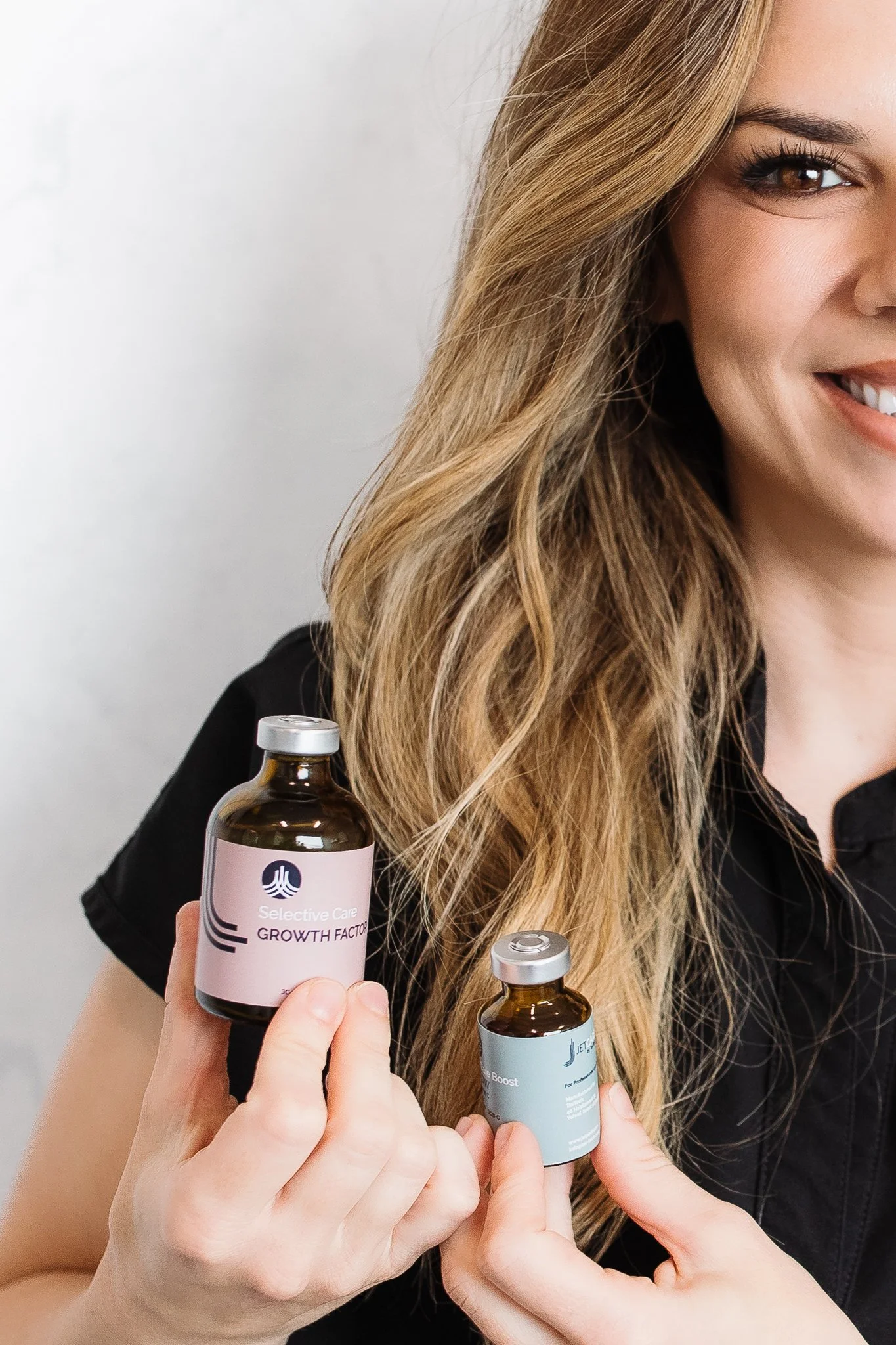 A woman with long blonde hair holding two small bottles of health supplements, one labeled 'Selective Care Growth Formula' and the other 'Jet Peel Boost,' smiling and wearing a black shirt.