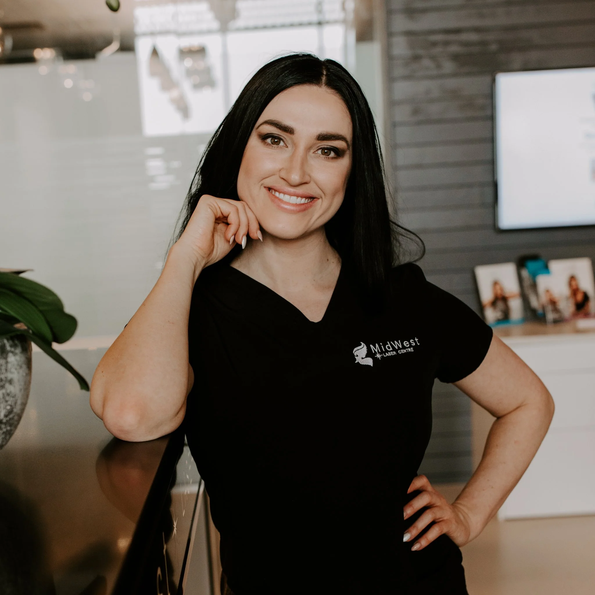 A woman with black hair wearing a black t-shirt with 'MidWest Laser Centre' logo, standing inside an office, smiling and resting her chin on her hand.