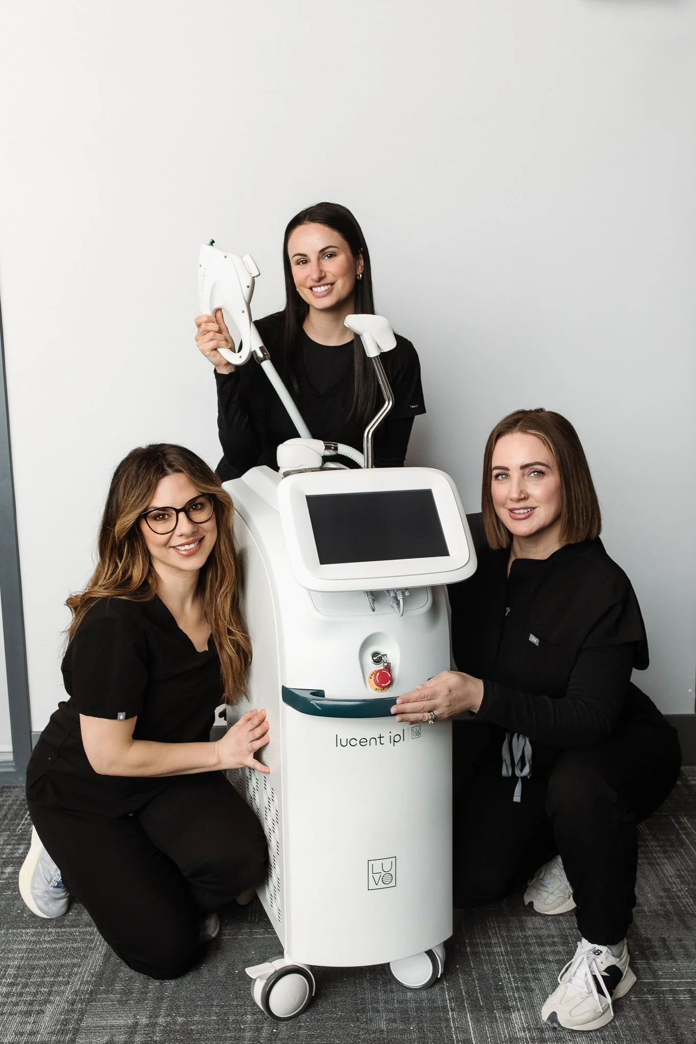 Four women in black uniforms gathered around a medical device with a screen, holding various medical tools, in a clinical setting with plain white and grey walls.