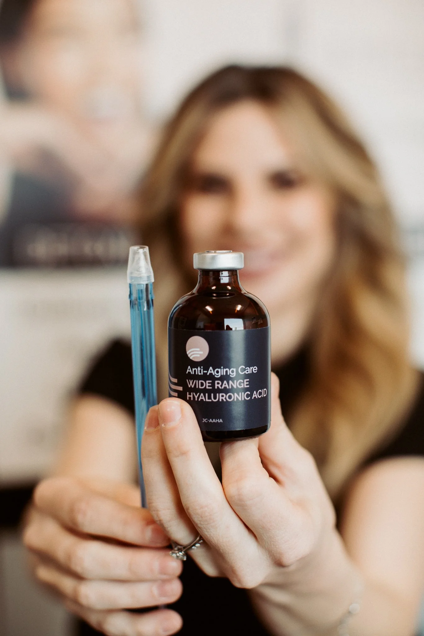 A woman with long, light brown hair holding a small brown bottle labeled 'Anti-Aging Care Wide Range Hyaluronic Acid' with a dropper, blurred background.