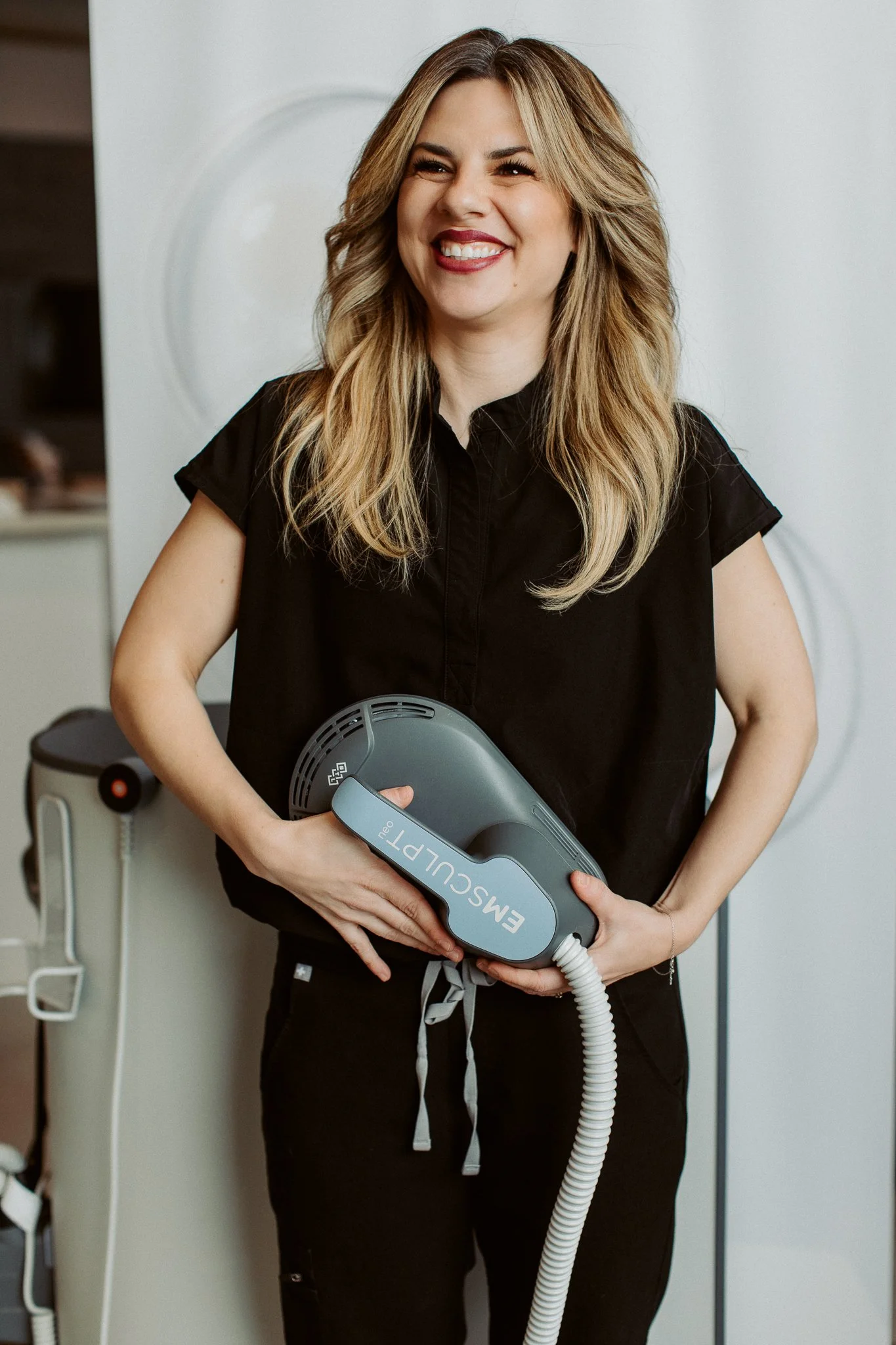 A woman smiling and holding a medical device labeled 'EMSculpt Neo' in a clinical setting.
