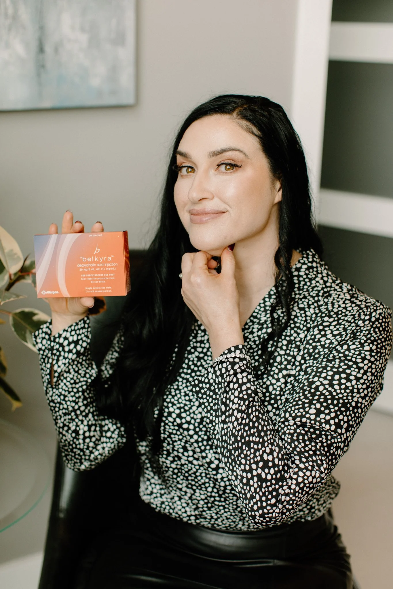 A woman with long black hair and light makeup, wearing a black and white polka-dot blouse, is sitting and smiling while holding a box of Belkyra deoxycholic acid injection.