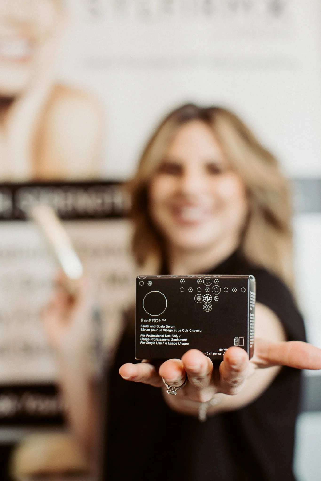 A woman taking a selfie in a mirror while holding a box of skincare serum, smiling.