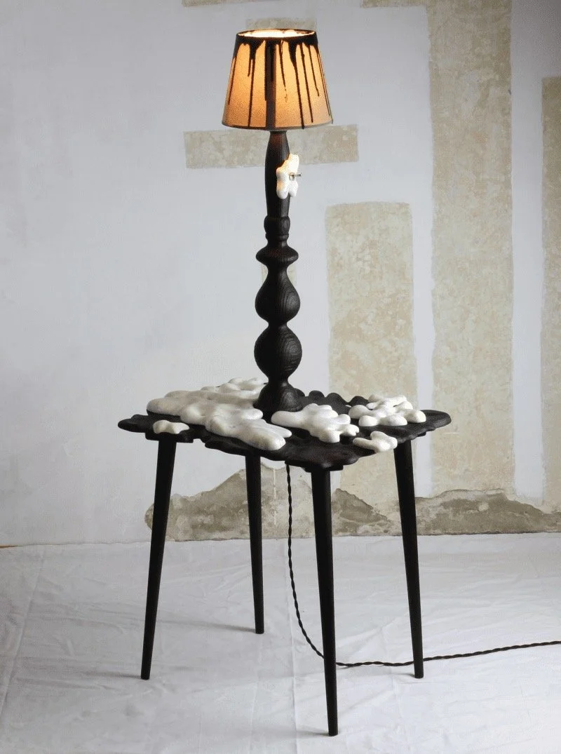 A black wooden table with a unique design, topped with a tall black lamp with a beige lampshade, and a small white decorative object attached to the lamp. The table surface is covered with white irregular-shaped objects resembling foam or plaster. Th