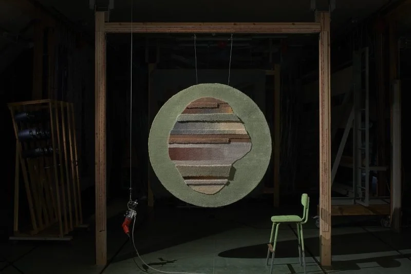 A round rug hangs in a wooden structure with a green chair underneath. The rug hangs from a wooden frame.