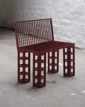 Red metal bench with grid design on the seat and backrest, placed against a gray brick wall on a concrete floor.