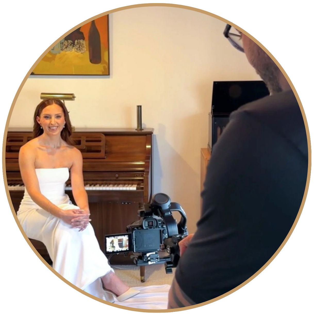 A woman in a white strapless dress sitting on a piano bench, smiling for a photo or video being taken by a person with a camera, in a room with artwork on the wall.