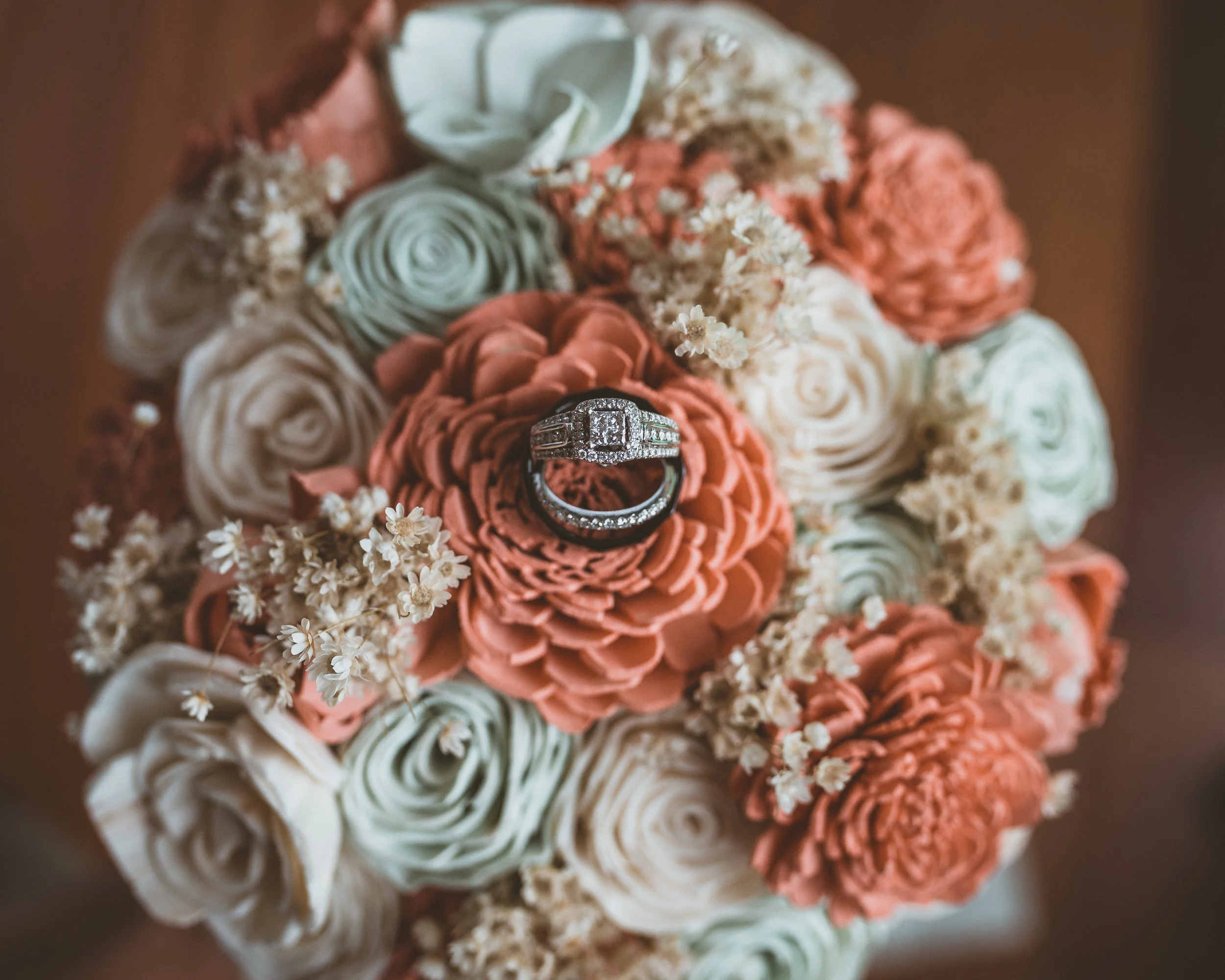 Close-up of handmade roses crochet flowers in cream, peach, and sage green colors, with a diamond engagement ring placed on a peach flower, surrounded by small white flowers.