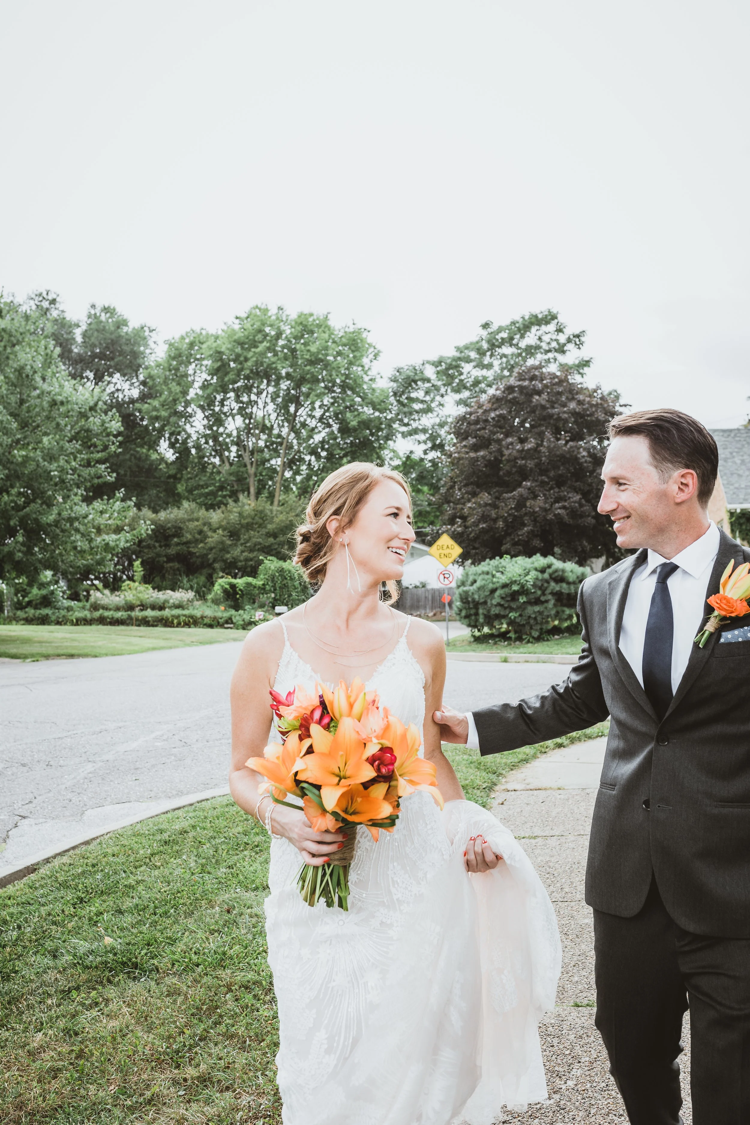 A bride and groom are smiling at each other outdoors, with the bride holding a bouquet of orange and red flowers, and the groom reaching out to hold her hand. There are trees and a yellow