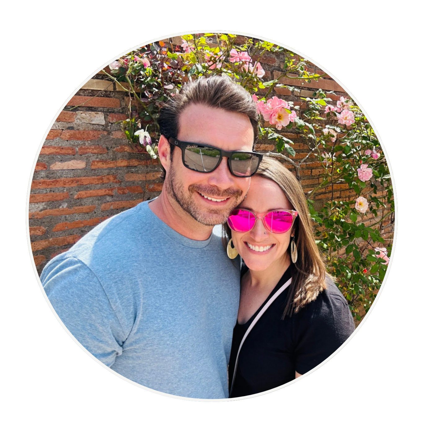 A smiling man and woman standing close together outdoors in front of a brick wall and pink flowering bushes, both wearing sunglasses.
