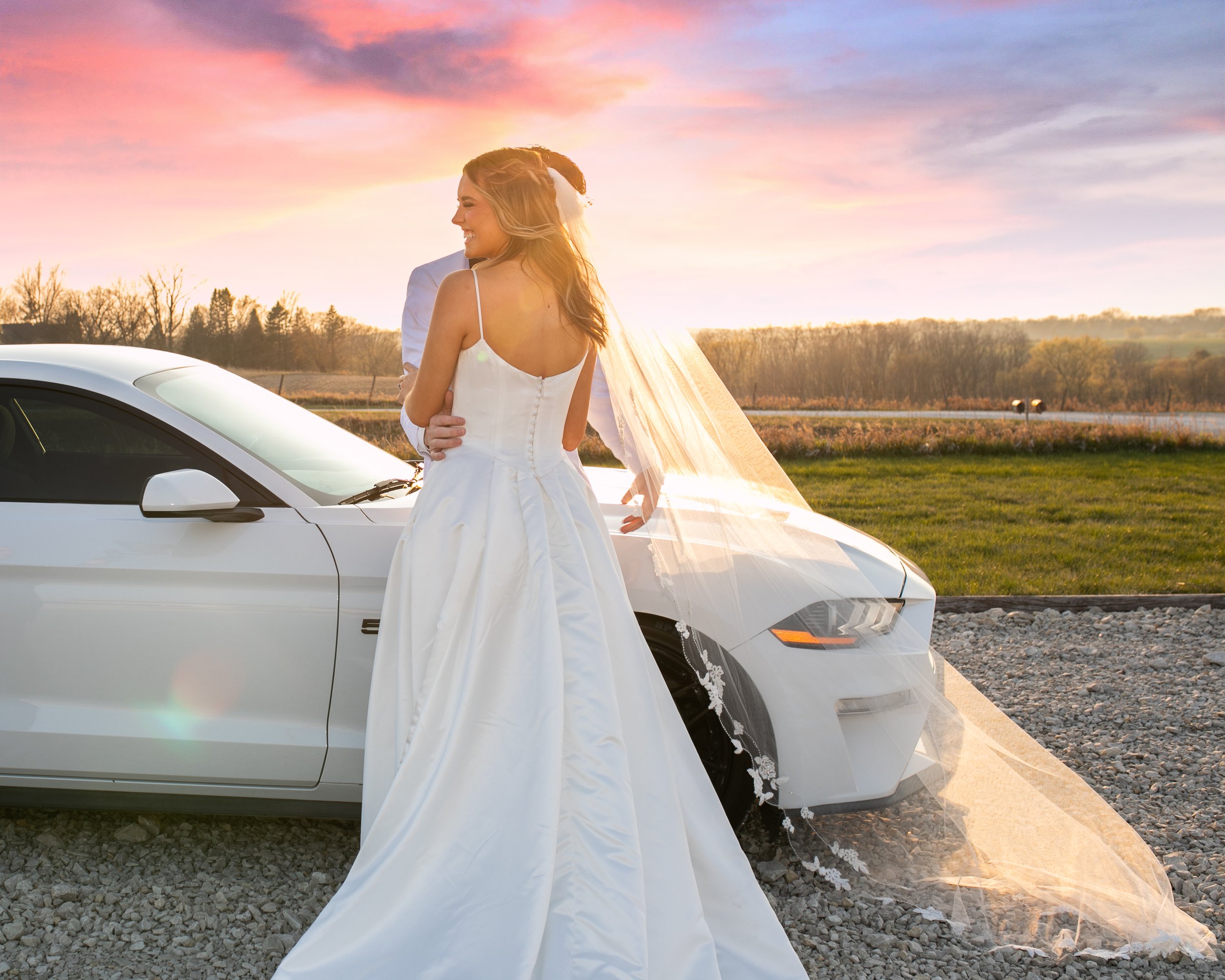 A happy bride in a white wedding dress with spaghetti straps, standing next to a white car with a sunset sky in the background.