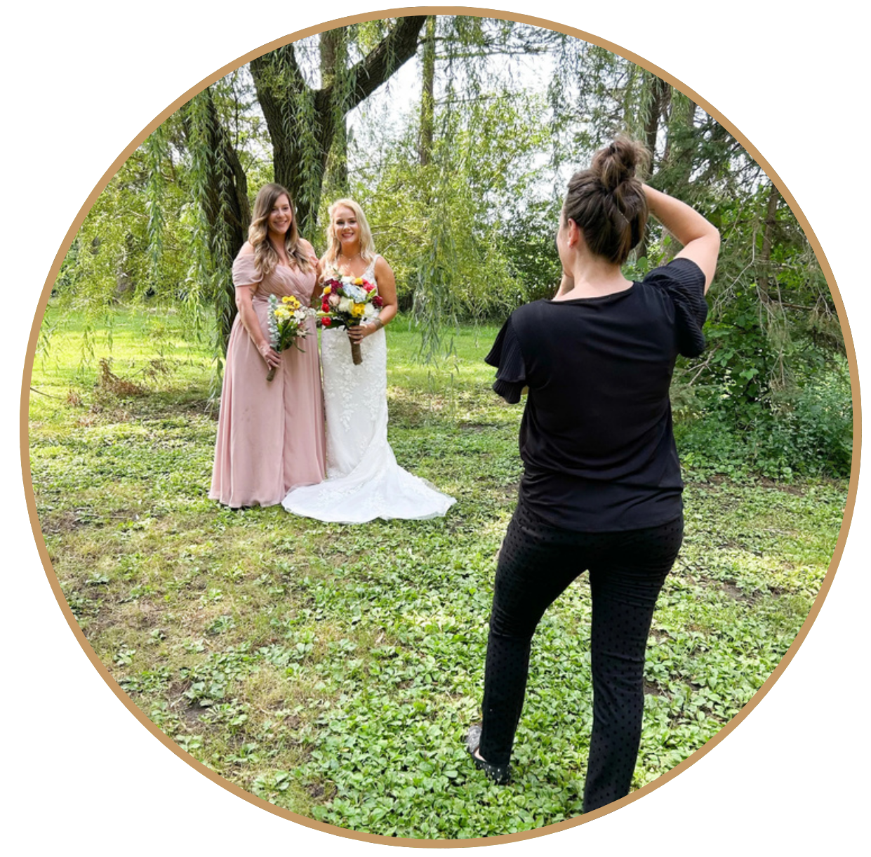 Two women in wedding dresses holding bouquets posing outdoors for a photo taken by a woman in black clothing under a tree with green foliage.