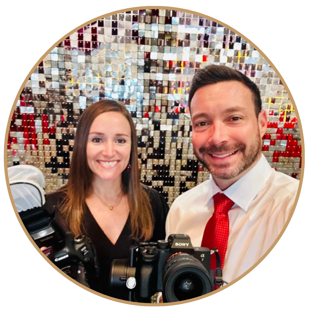 A smiling man and woman taking a selfie in front of a mosaic wall with reflective, mirror-like tiles. The man is wearing a white shirt and red tie, and the woman is in a black top. They are holding cameras.