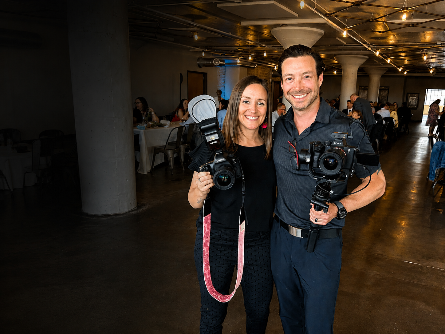 A man and woman smiling and holding professional cameras at an indoor event with tables and people in the background.