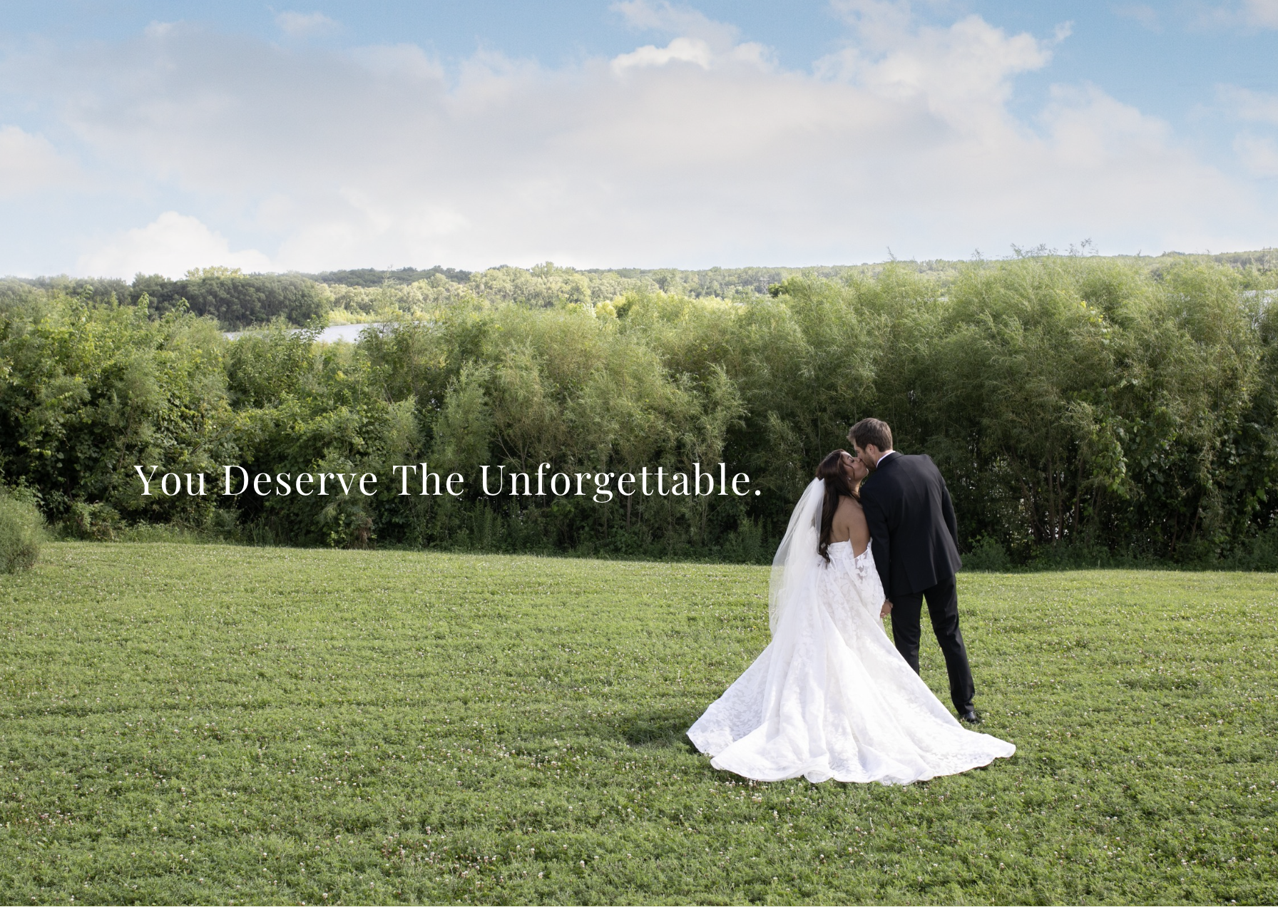 A newlywed couple sharing a kiss outdoors on a grassy field with trees and a blue sky in the background. The bride wears a white wedding dress and veil, and the groom is in a black suit. Overlaid text reads, 'You Deserve The Unforgettable.'