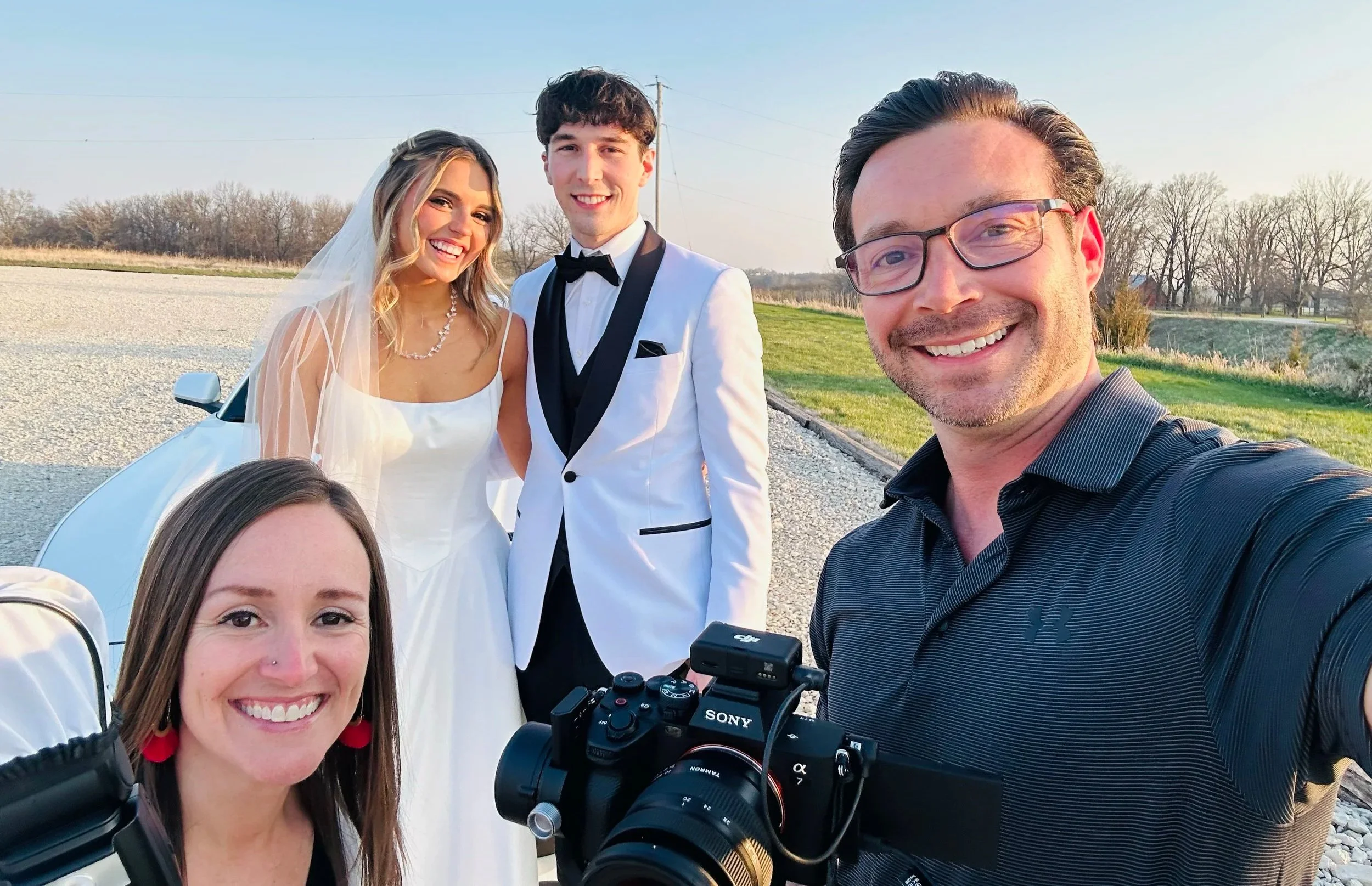 A group of five friends taking a selfie outdoors during sunset. The group includes a bride in a wedding dress and veil, a groom in a tuxedo, and three other friends, including a man with glasses holding a Sony camera, and two women smiling at the camera. The background is a rural landscape with trees and open fields.