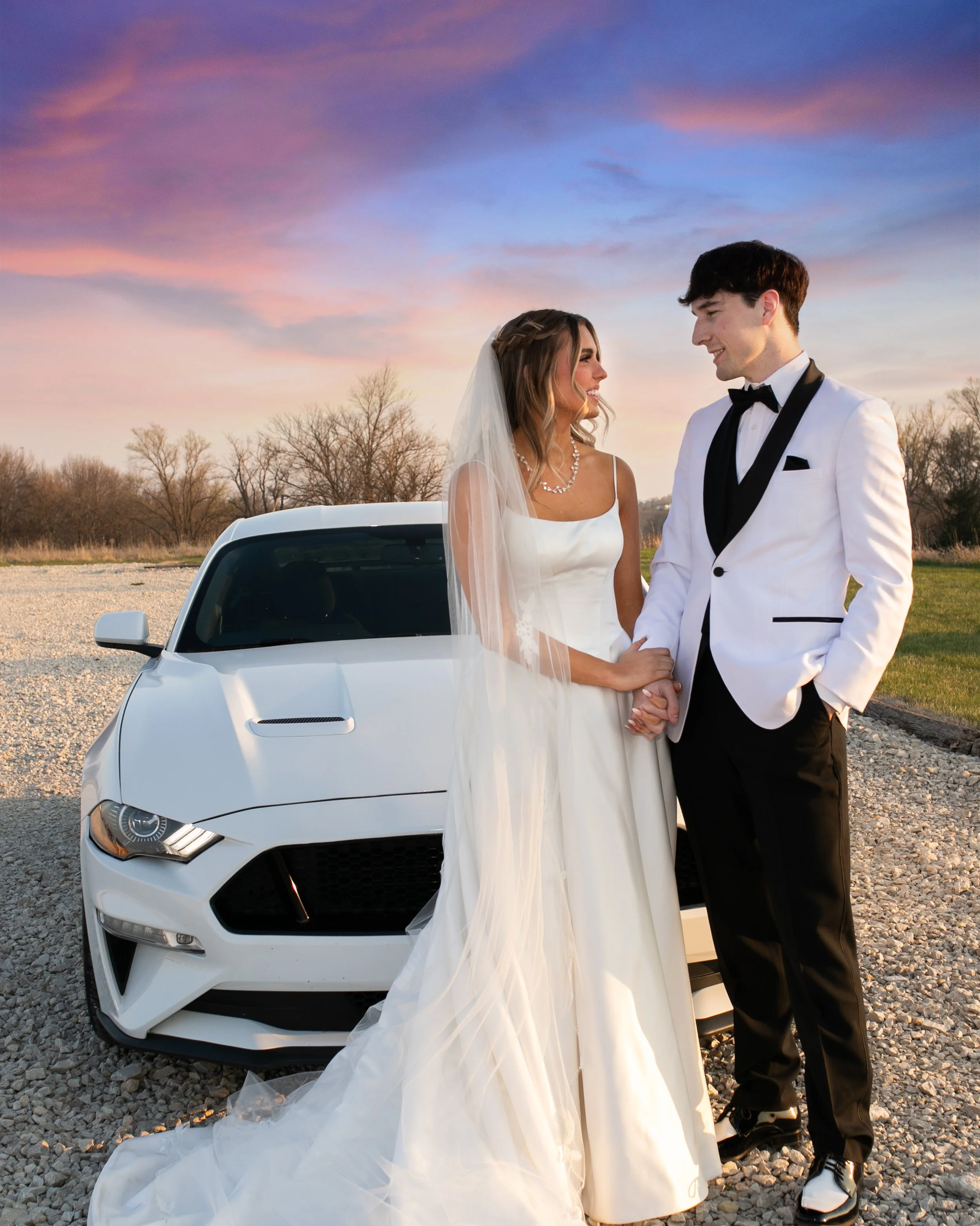 A bride and groom in wedding attire holding hands next to a white car outdoors during sunset, with trees and a sky with pink and purple clouds in the background.