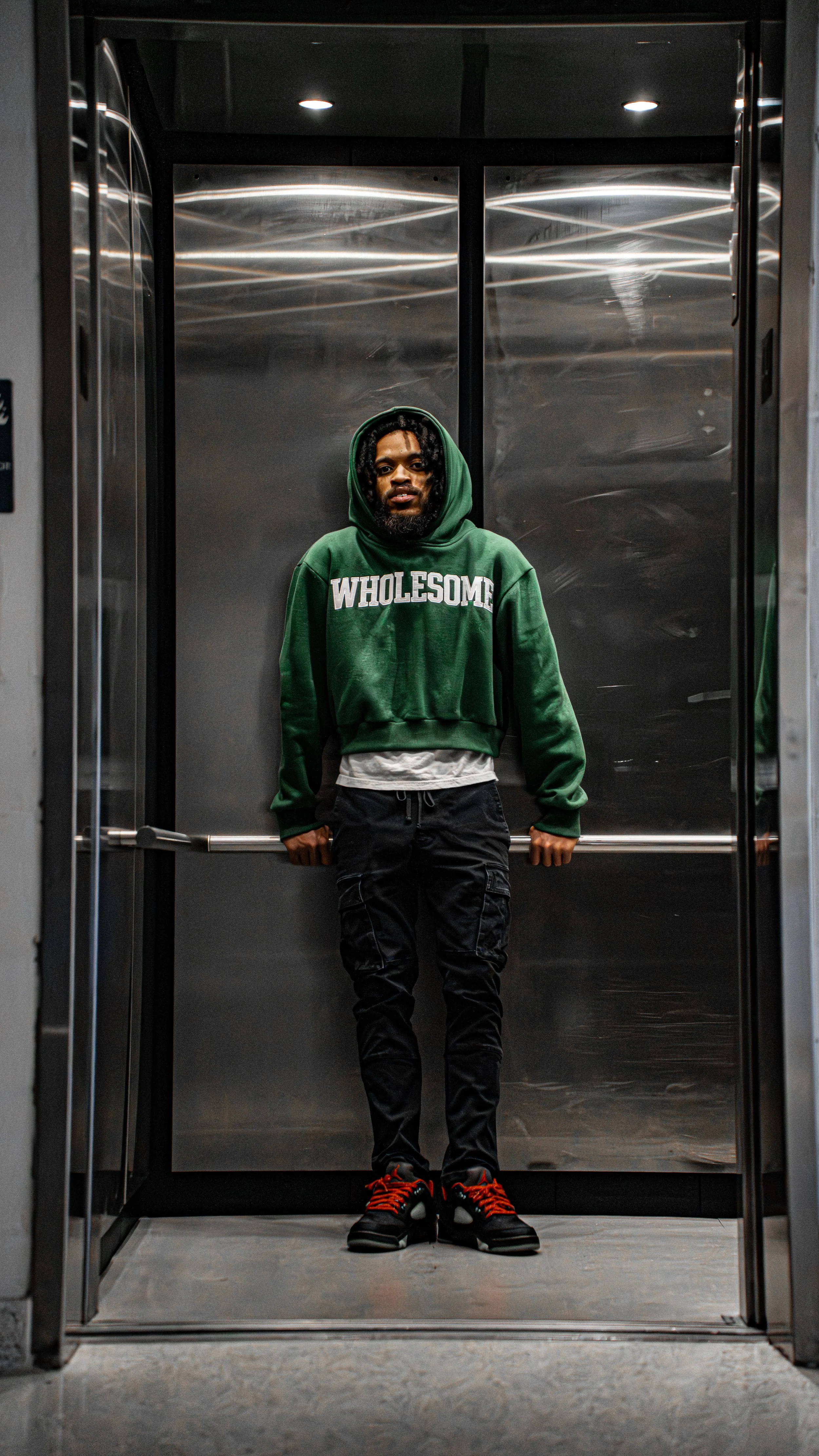 A man with a beard and curly hair stands inside an elevator, wearing a green hoodie with the word 'WHOLESOME' on it, black pants, and black sneakers with red shoelaces.