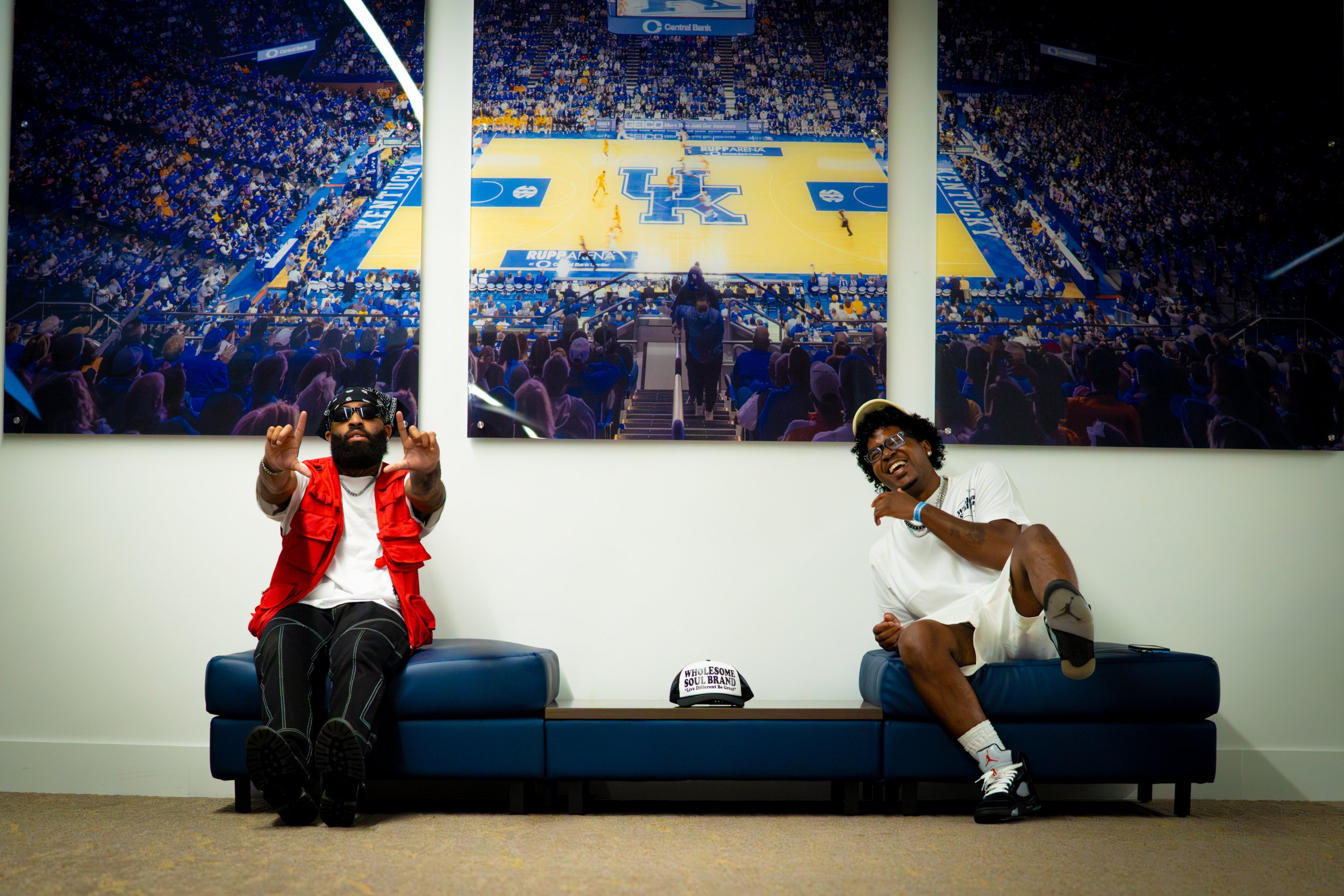 Two men sitting on a blue bench in front of a large sports arena map behind them. One man is wearing a red vest, black pants, sunglasses, and making a gesture with his hands. The other man is dressed in white, smiling, and raising his one knee. A cap