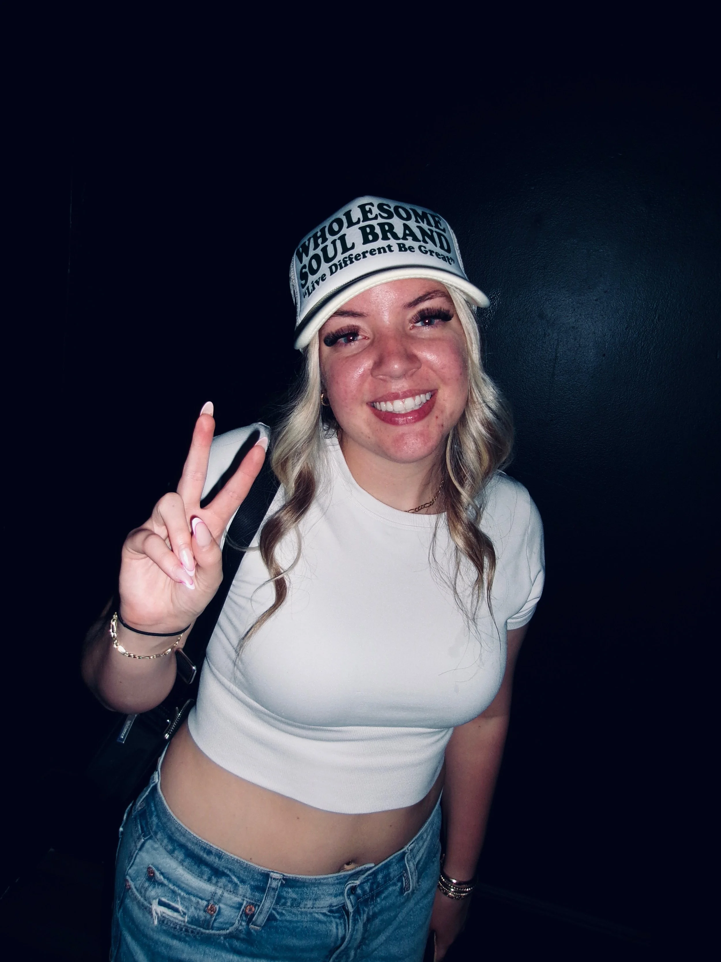 Young woman smiling and holding up a peace sign, wearing a white crop top, jeans, and a white cap with text.