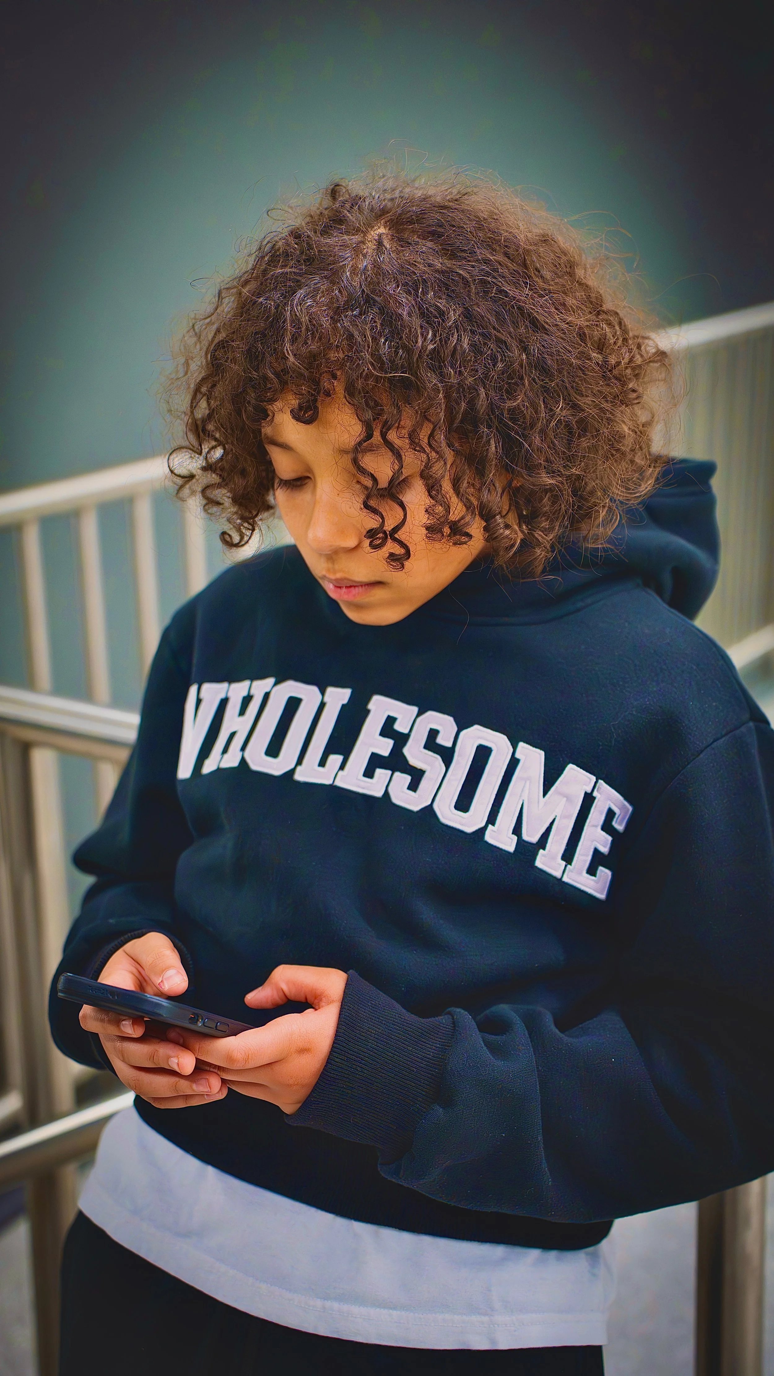 A young woman with curly hair wearing a navy hoodie with the word 'WOLESOME' on it, standing outdoors, looking at her smartphone.