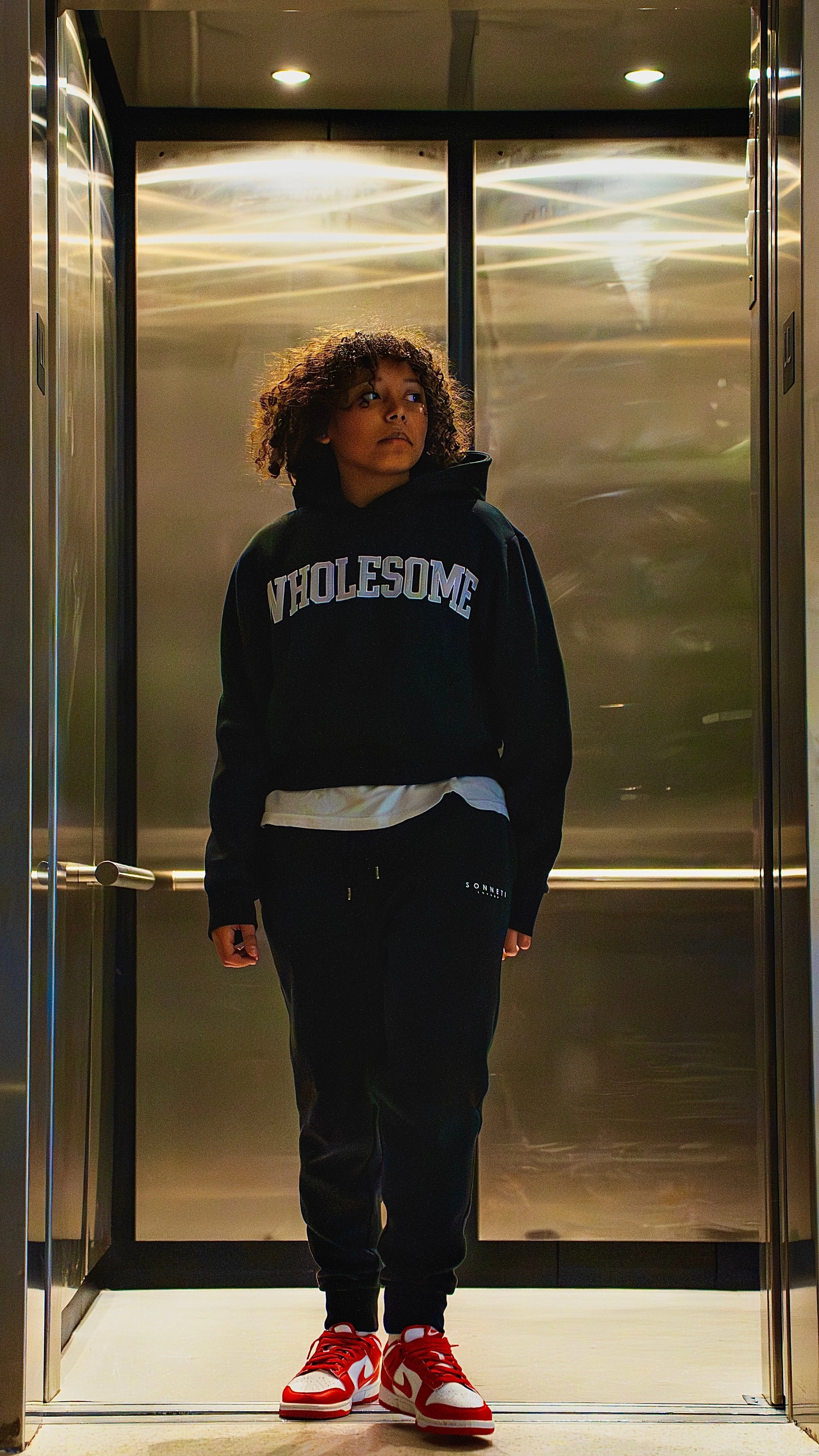 A young person with curly hair standing in an elevator, wearing a black hoodie with the word 'WHOLESOME' and red and white sneakers.