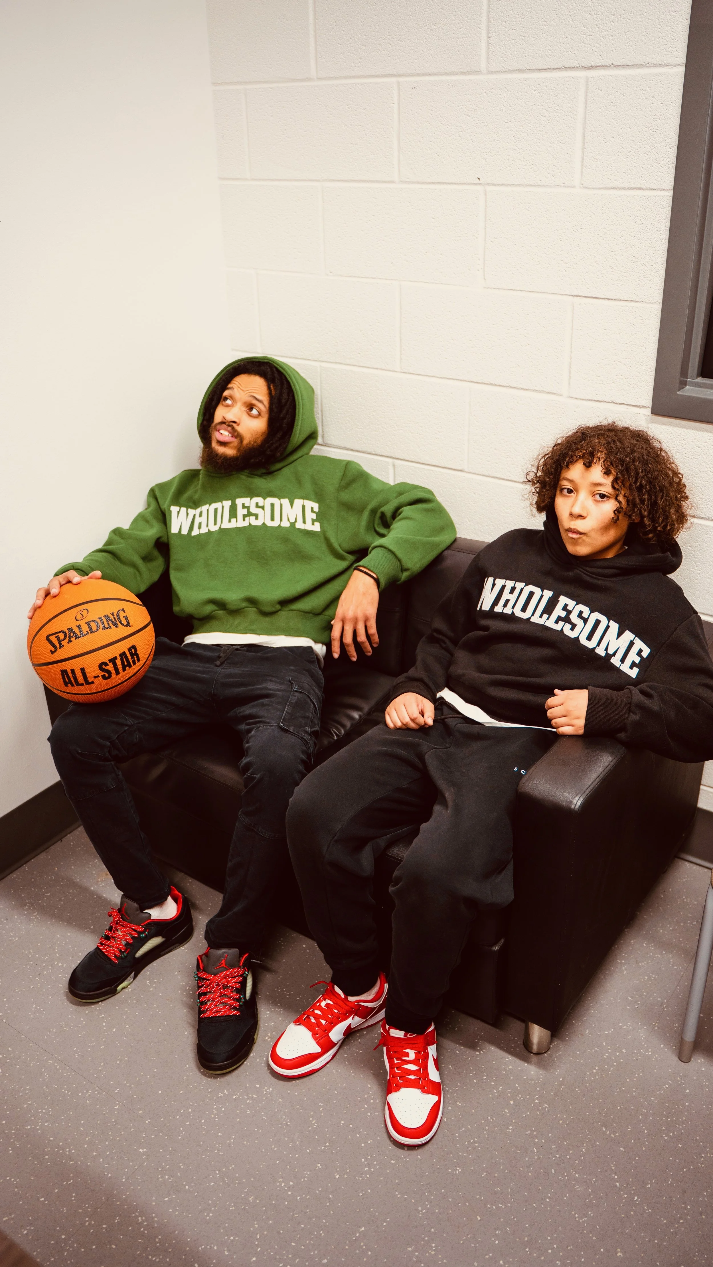 Two young men sitting on a black leather couch against a white cinder block wall. One is holding a basketball, and both are wearing hoodies that say 'WHOLESOME'. The man on the left wears a green hoodie, black pants, and sneakers with red laces. The 