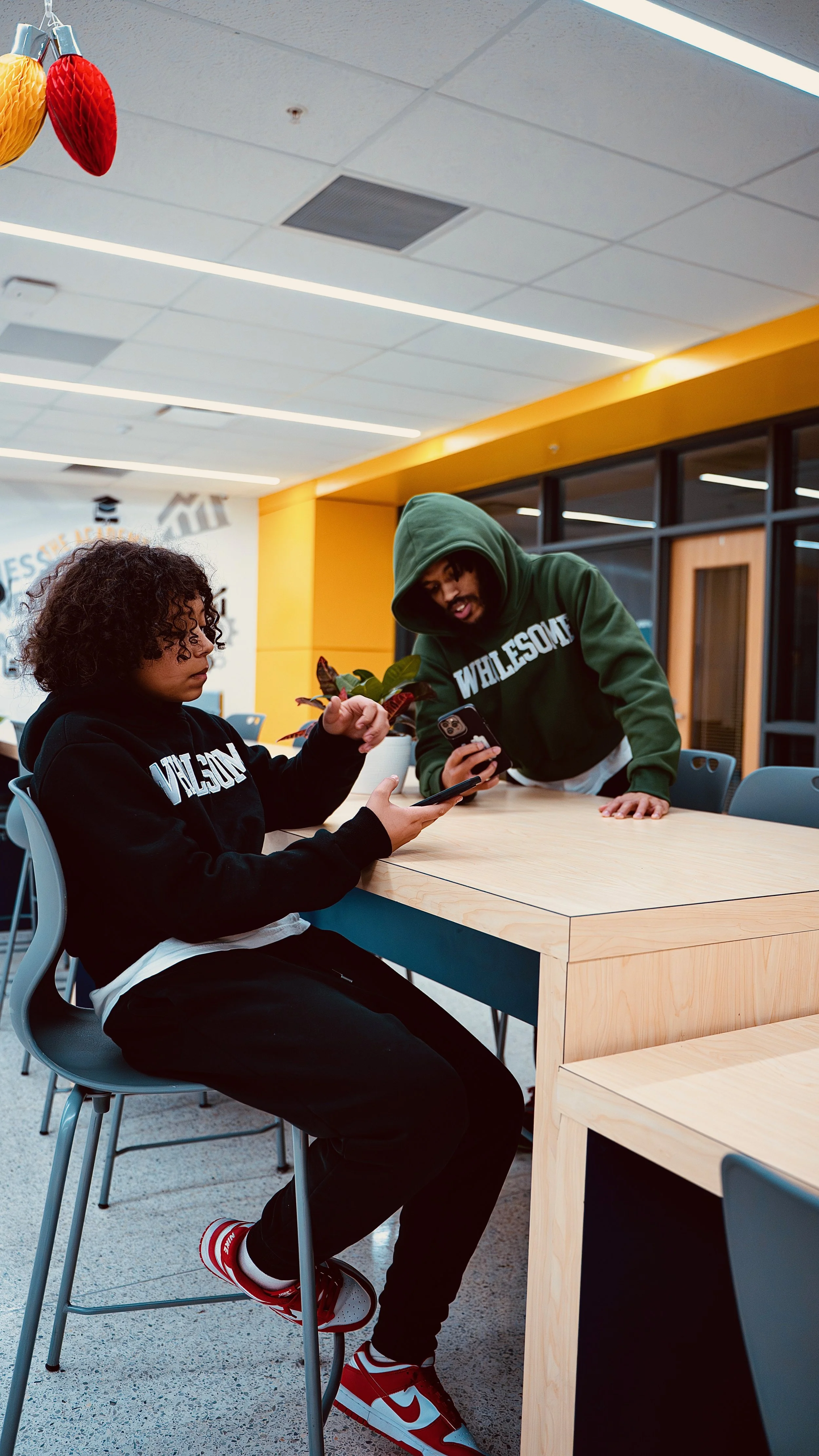 Two young people sitting at a light wooden table, looking at a smartphone together inside a modern, brightly lit room with yellow walls and large windows. Both are wearing hoodies with the word 'WHolesome' printed on them.