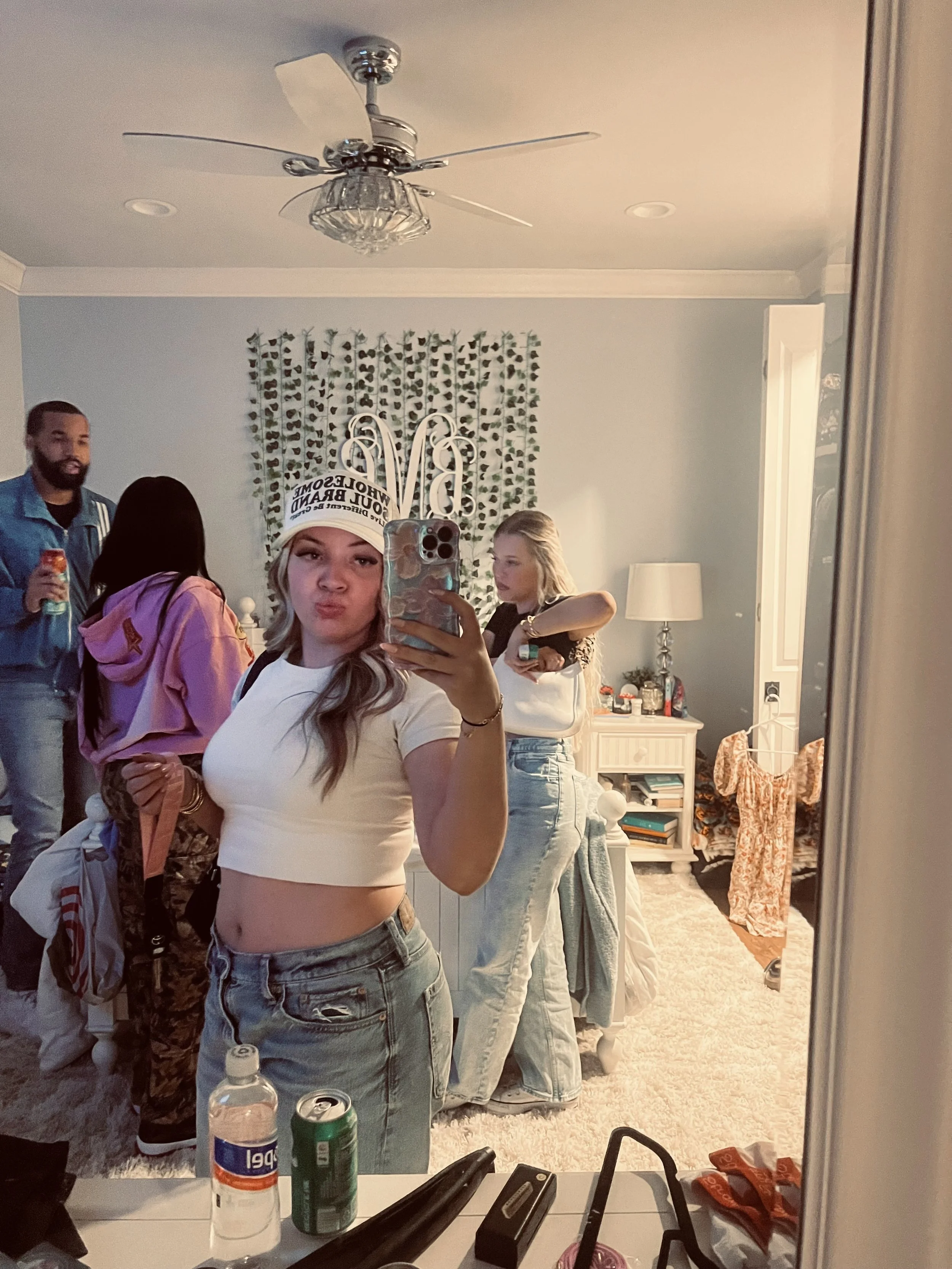 A young woman taking a selfie in a mirror at a party or gathering, wearing a white crop top, jeans, and a birthday hat that says 'World's Most Awesome Soul Bear.' Four other people are in the background, one man holding a drink, two women chatting, a