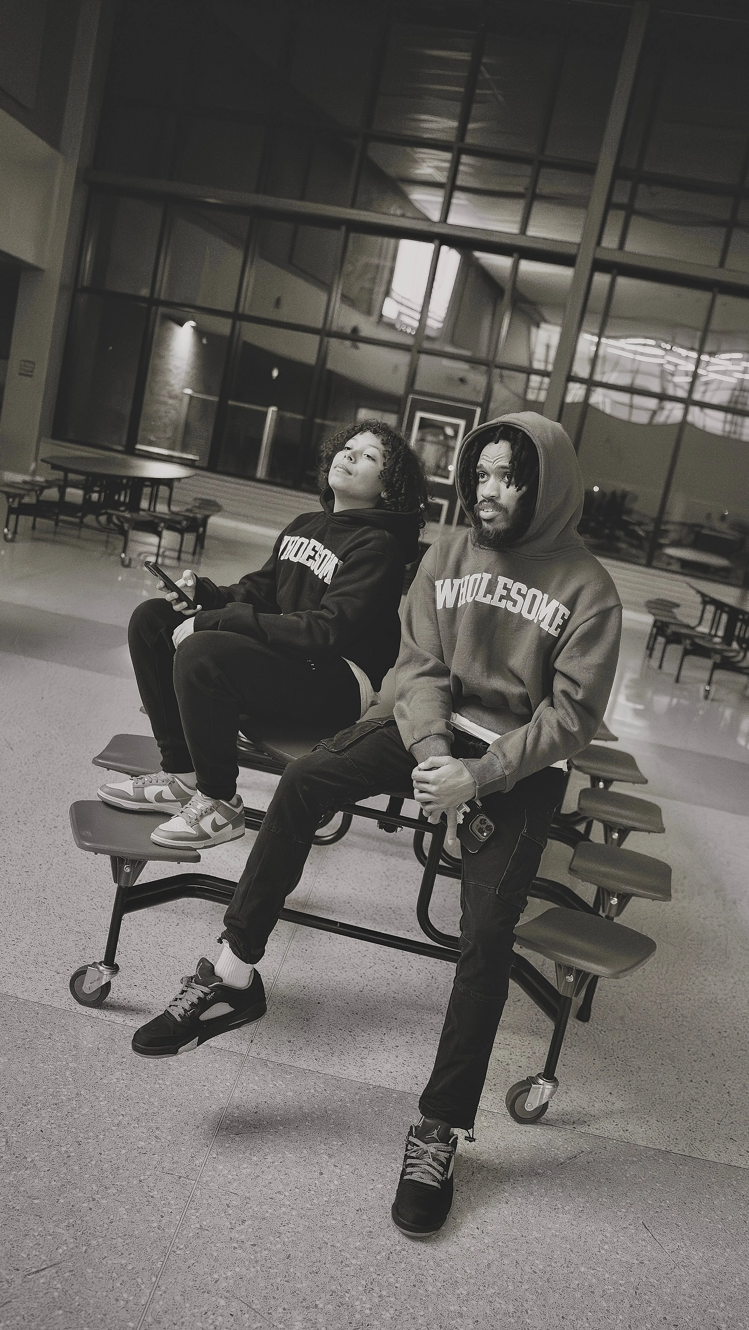 Two young men sit on a movable bench in an indoor space, one on the left holding a phone and the other on the right with his hands clasped, both wearing hoodies with 'WIDESOME' printed on them. Behind them is a large glass wall with reflections, and 