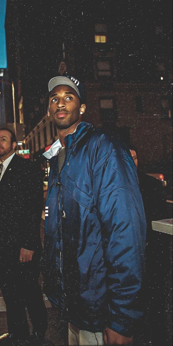 A young man wearing a blue jacket and a baseball cap walking at night on a city street, with a tall building and another person visible in the background.