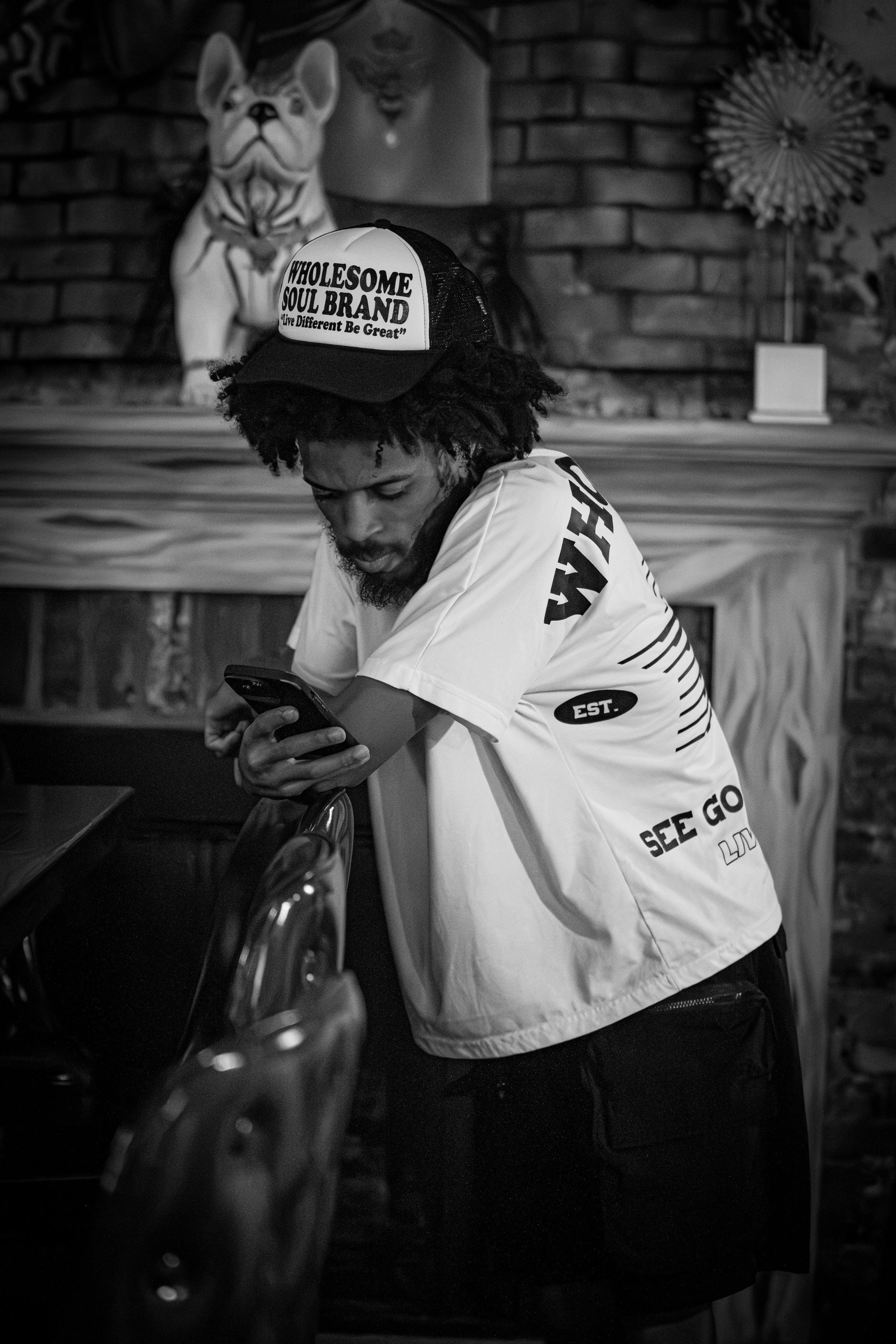 Man in a white t-shirt and trucker hat looking at his phone, standing near a table in a room with brick wall and decorative items.