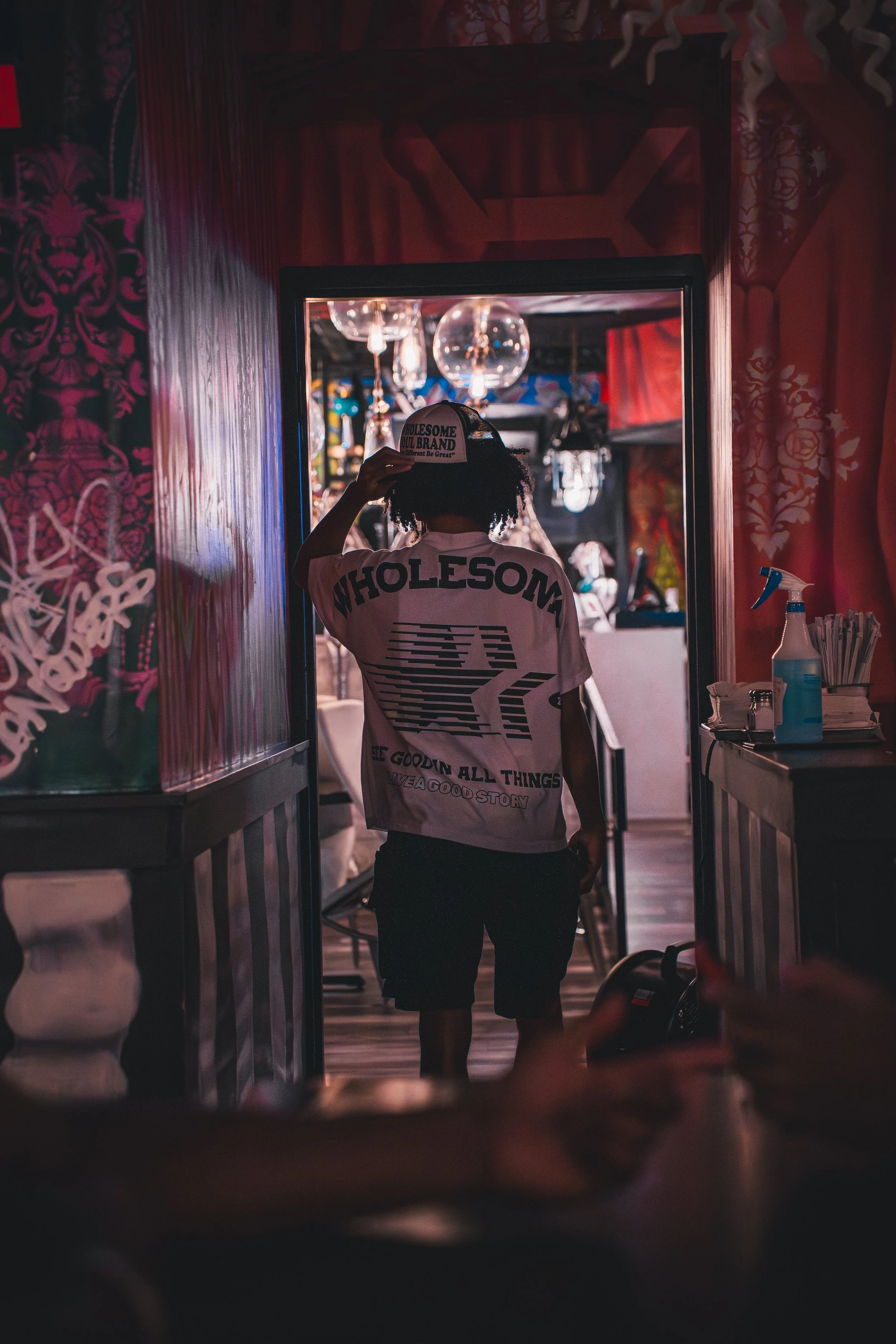 A person standing in front of a mirror in a colorful, decorated room, wearing a white t-shirt with "WHOLESOME" written on the back and a cap, adjusting the cap's brim.