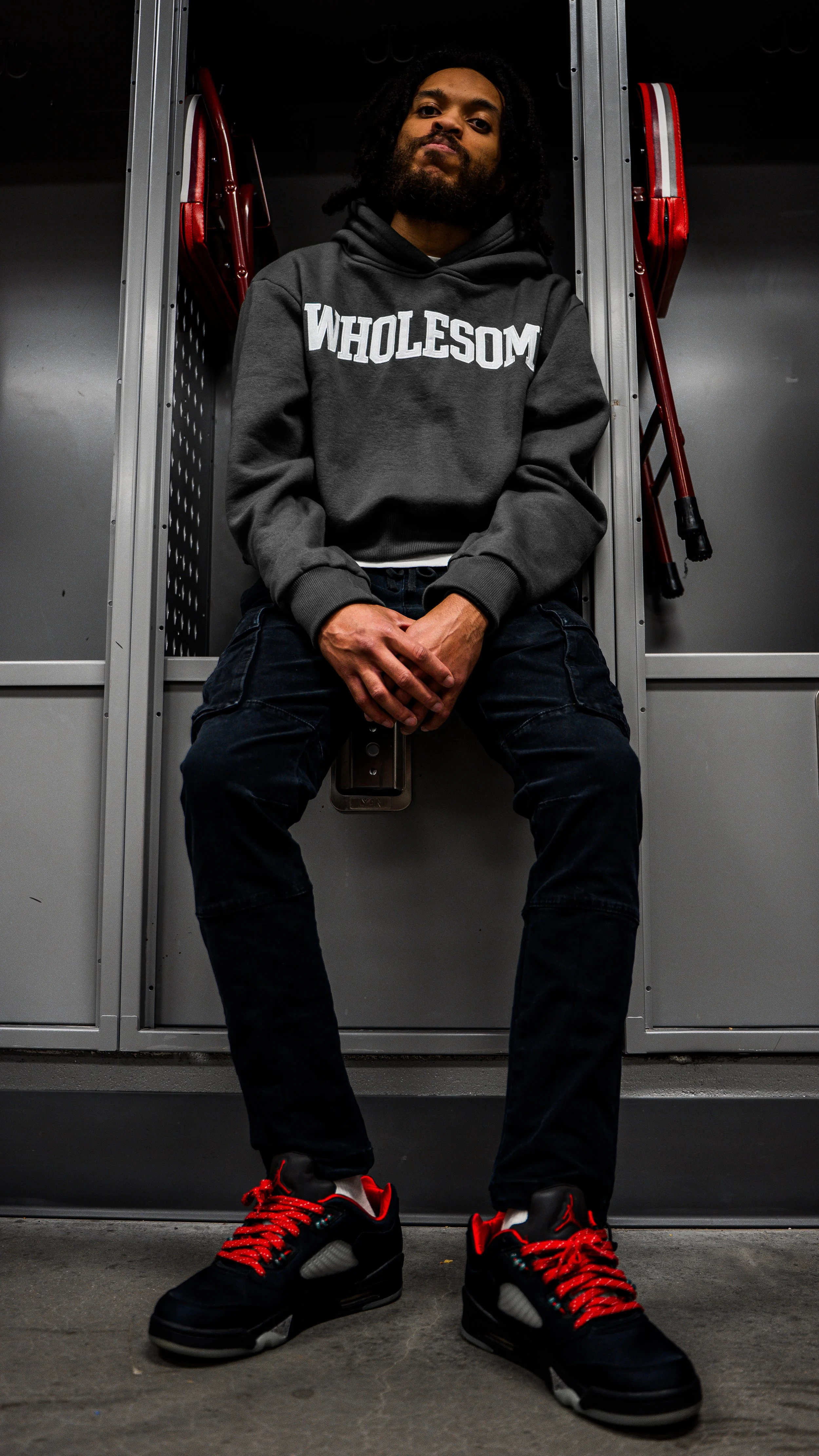 A man sitting in a locker room wearing a gray hoodie with 'WHOLESOM' printed on it, black pants, and black and red sneakers, with red folding chairs behind him.